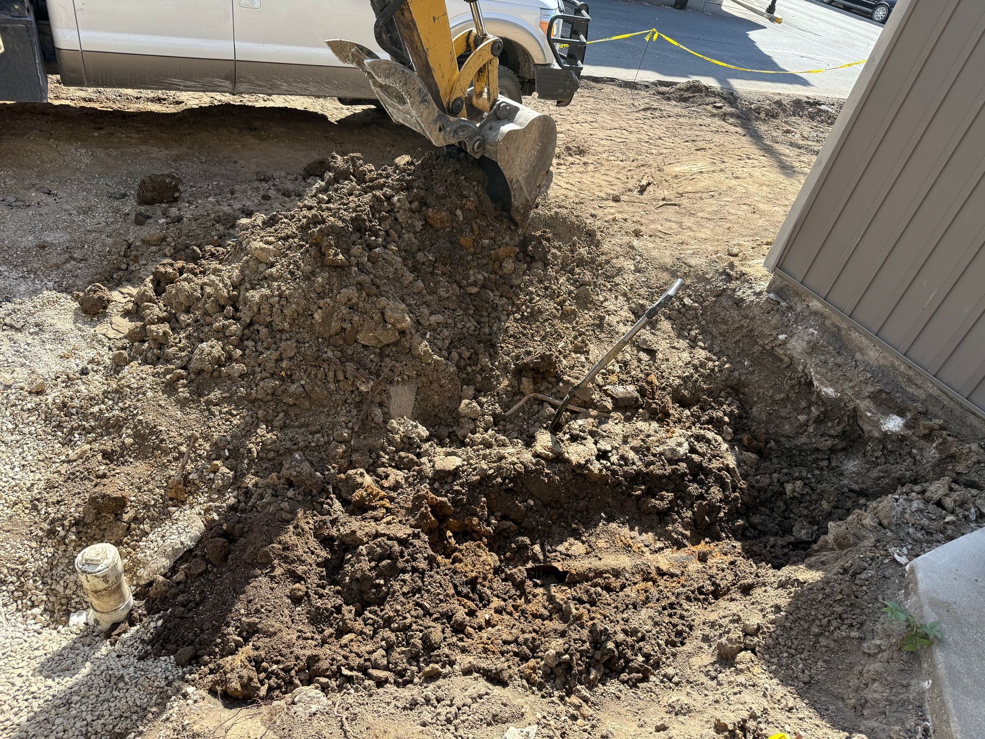 Excavator digging a large dirt trench beside a building and a concrete sidewalk