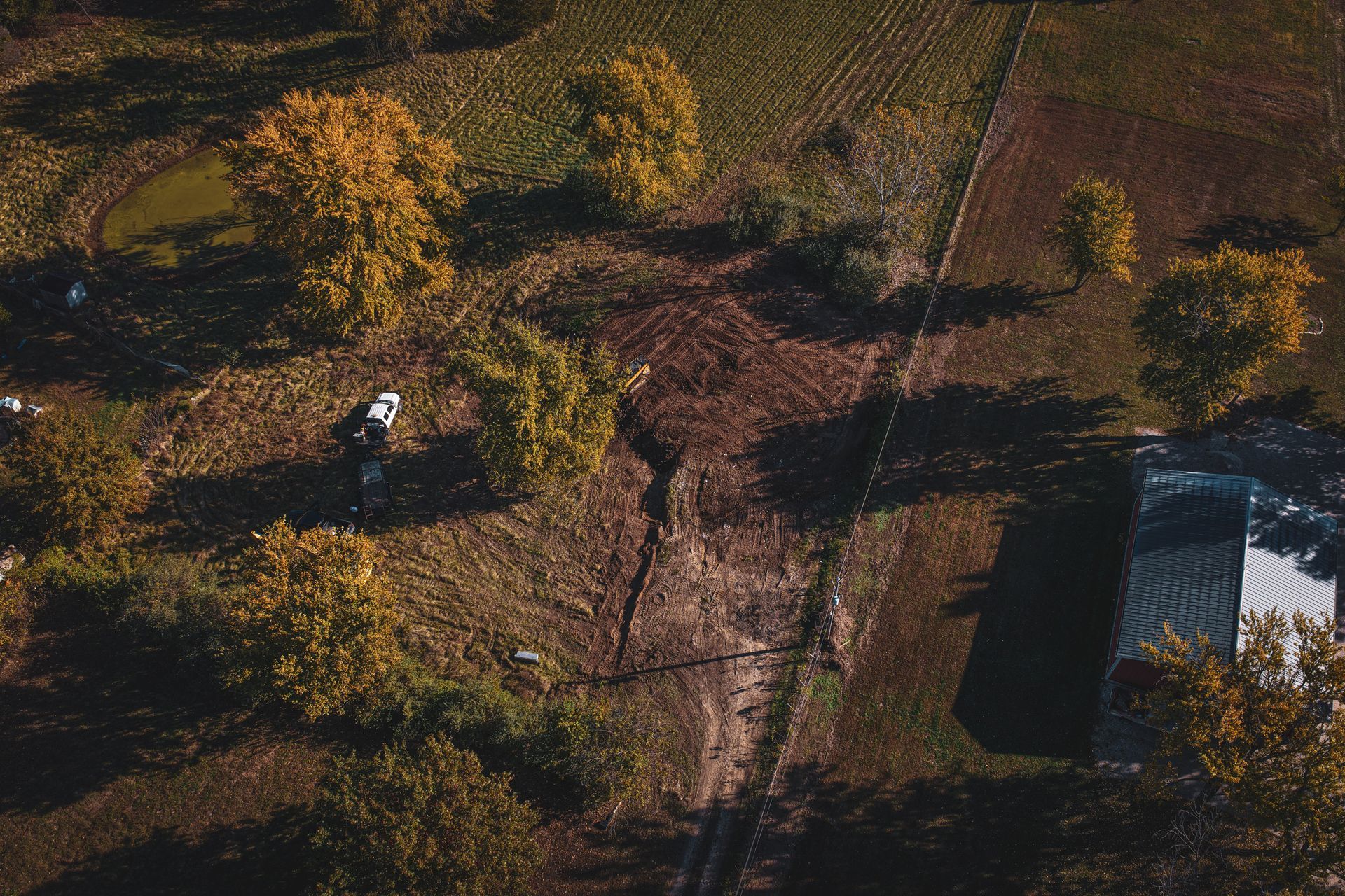 Aerial view of a person walking near trees and a small dark-roofed building in a sunlit field