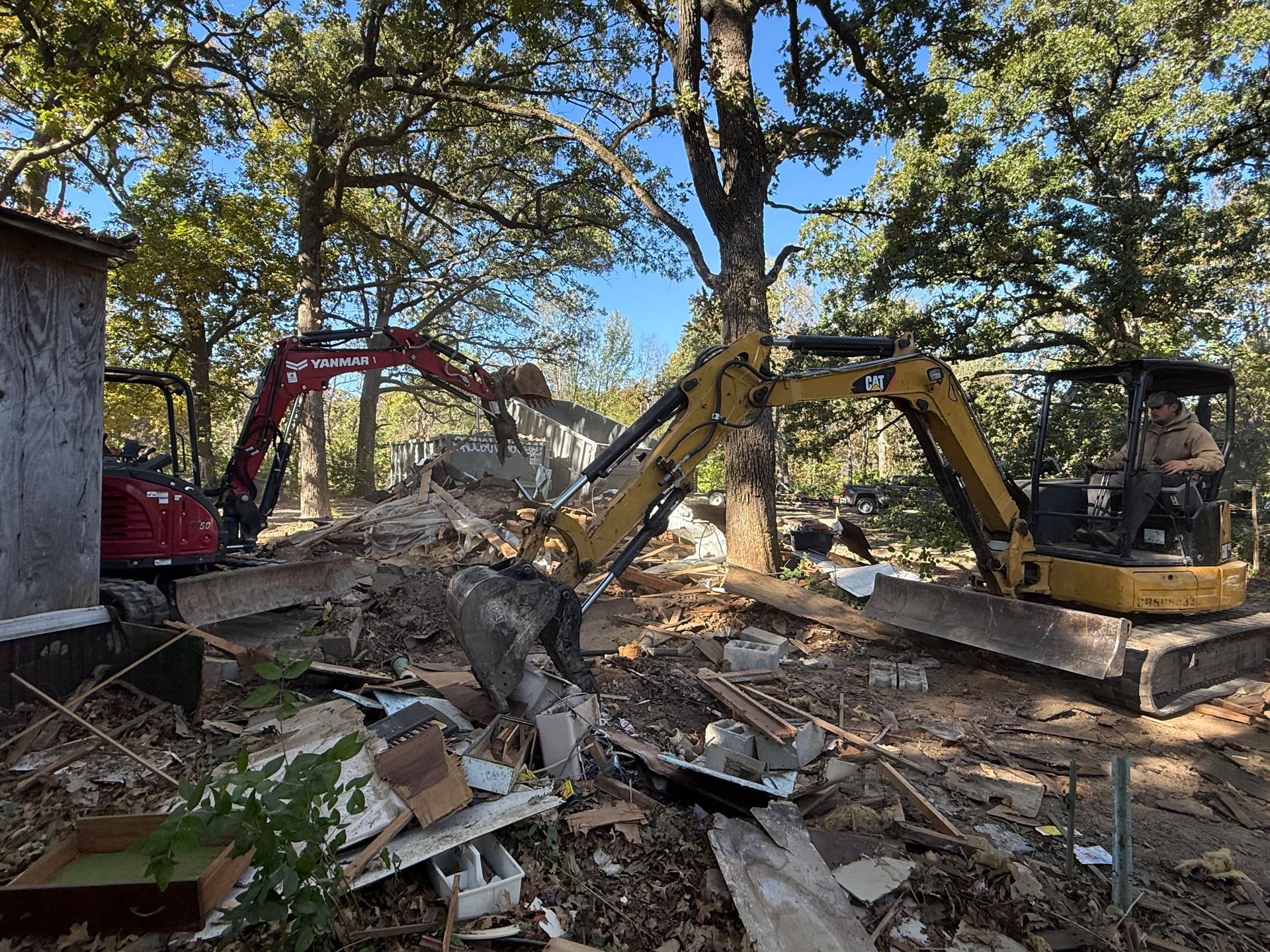 Two excavators clearing rubble at a demolition site beside a small building and trees
