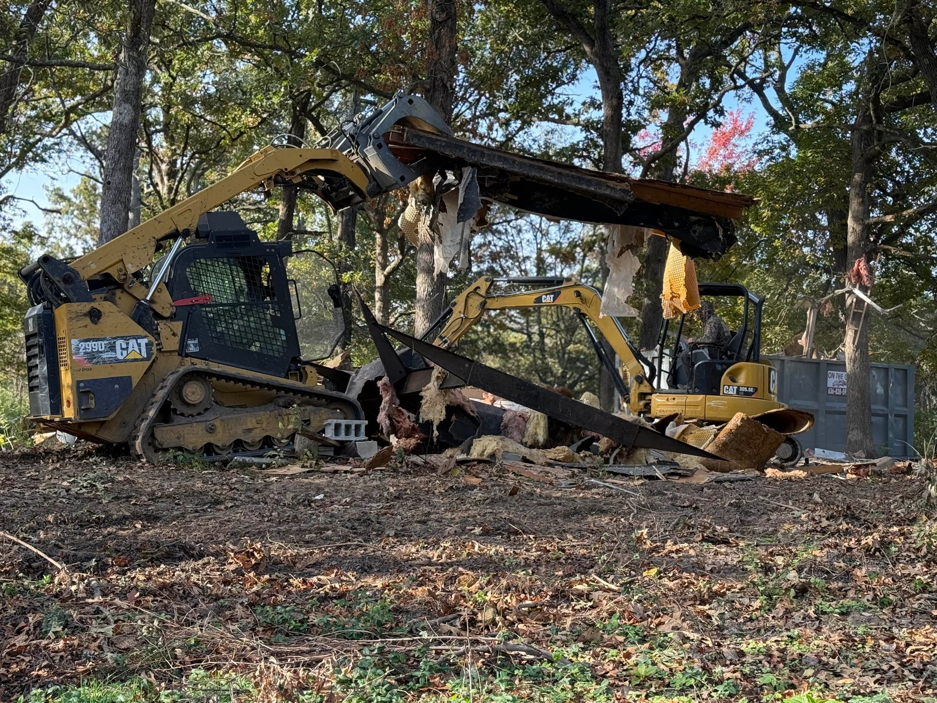 Yellow excavator tearing down a structure in a wooded area amid debris and trees