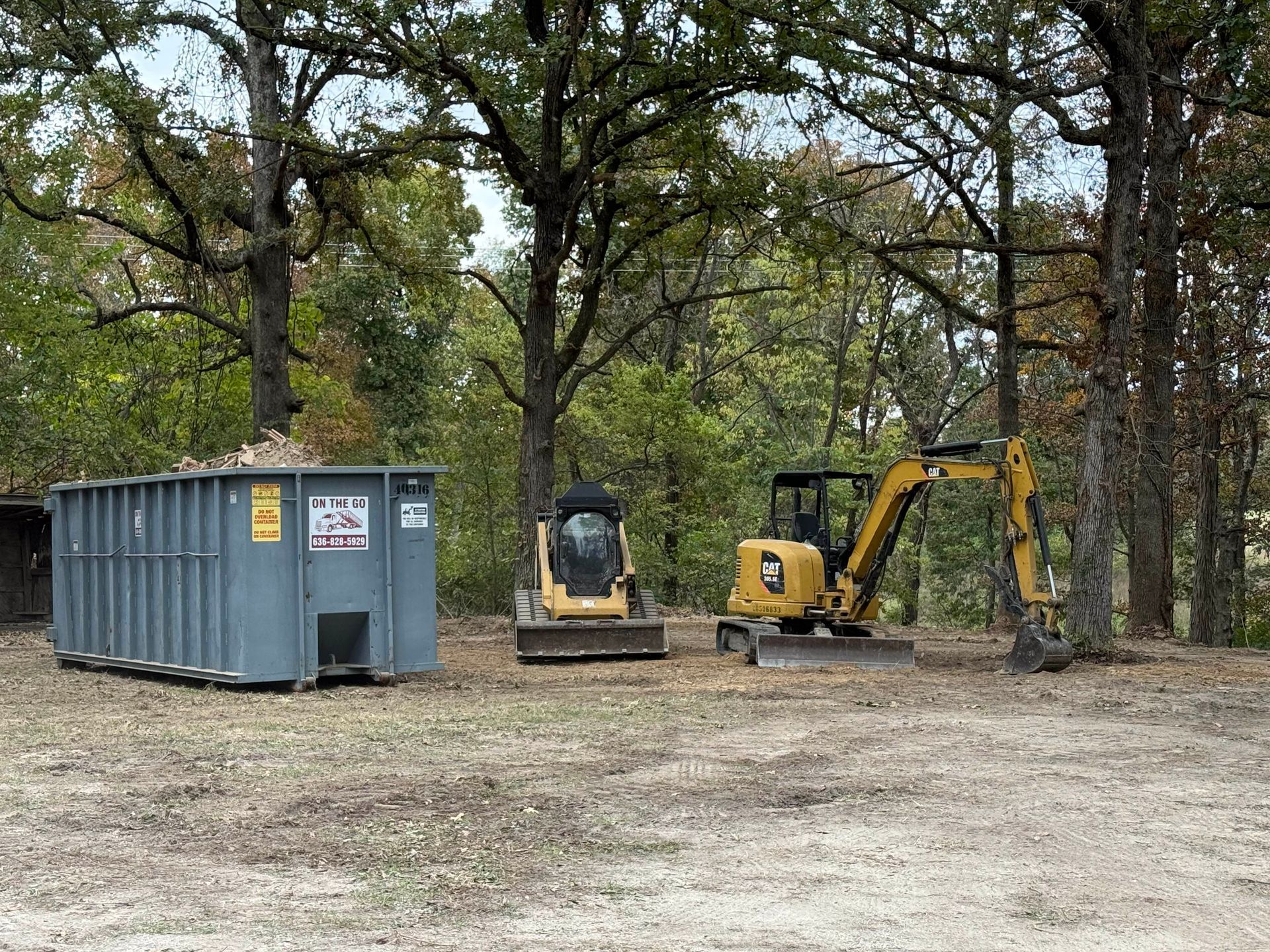 Yellow excavators beside a gray dumpster in a wooded dirt lot
