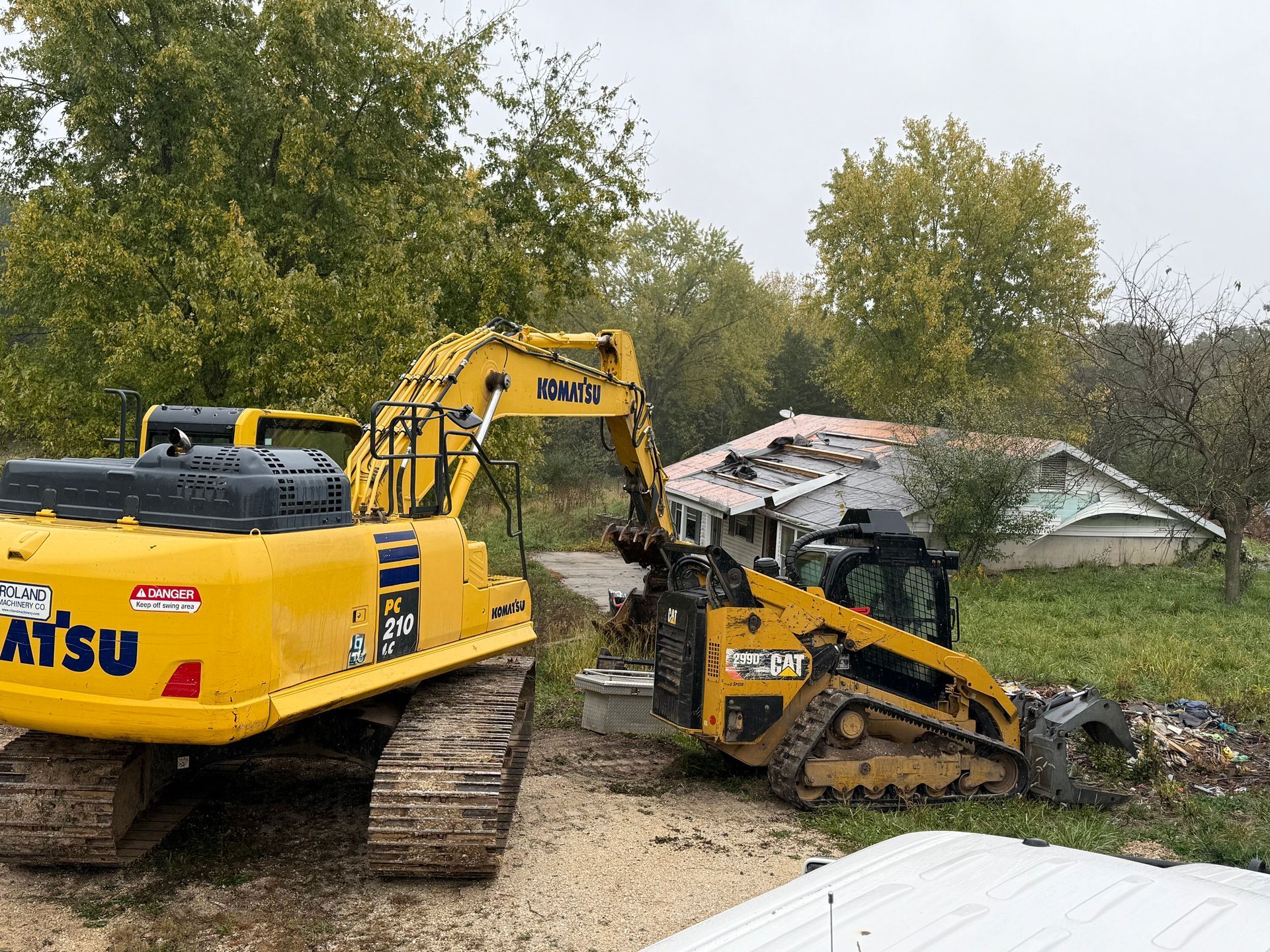 Yellow excavator and skid steer clearing debris beside a damaged house amid trees.