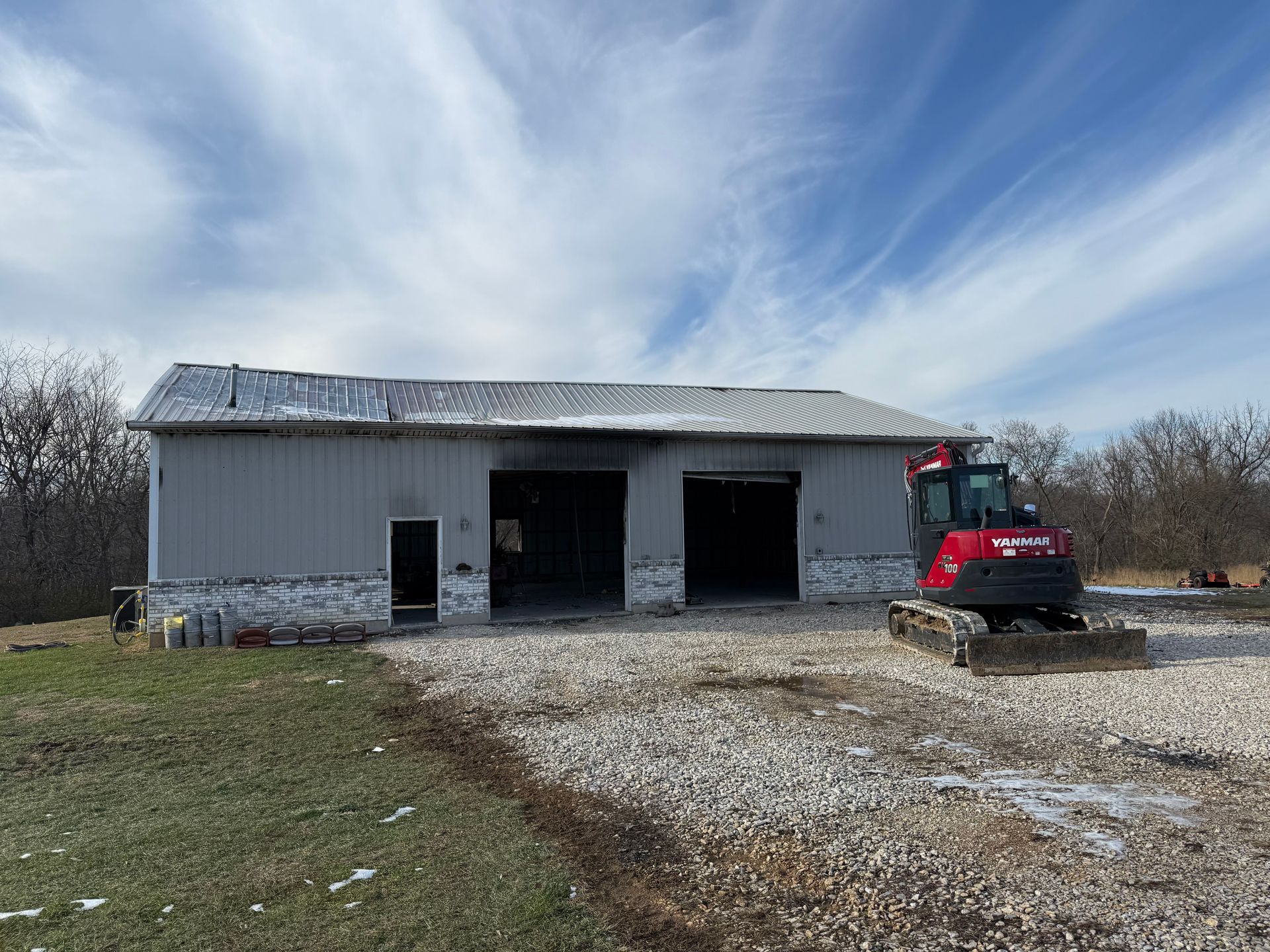 White barn with open bays and a red skid steer on a gravel driveway under a cloudy sky.