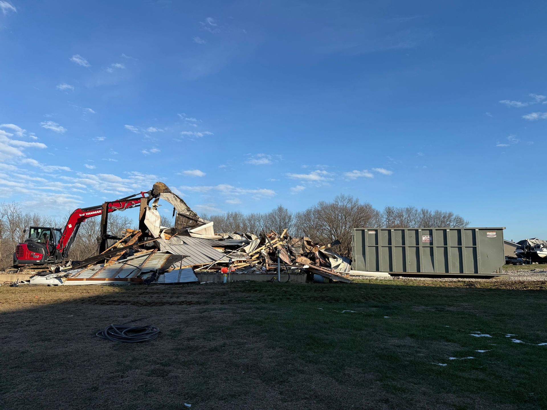 Excavator demolishing a pile of rubble beside a green dumpster under a blue sky