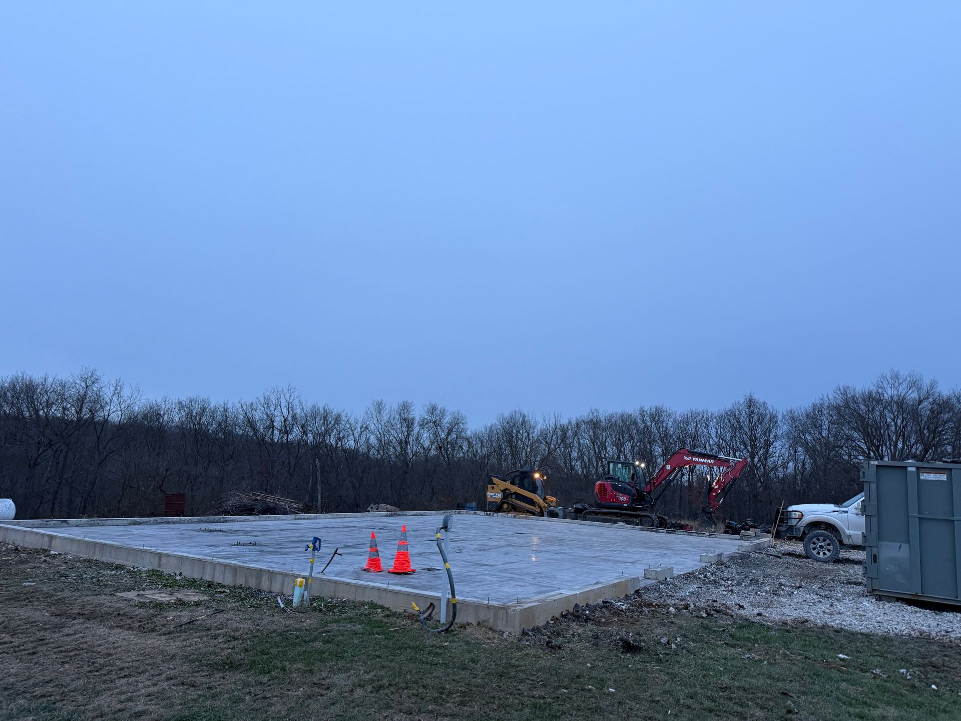 Construction site at dusk with excavator, red machinery, orange cones, and a white truck on a gravel lot