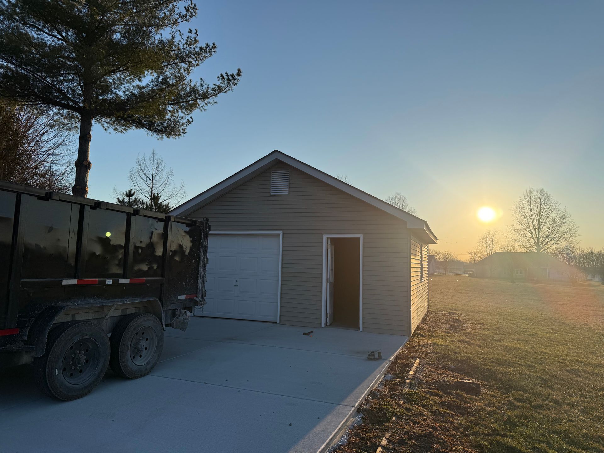 Sunlit driveway beside a beige garage and utility trailer at dusk, with trees and a hazy horizon.