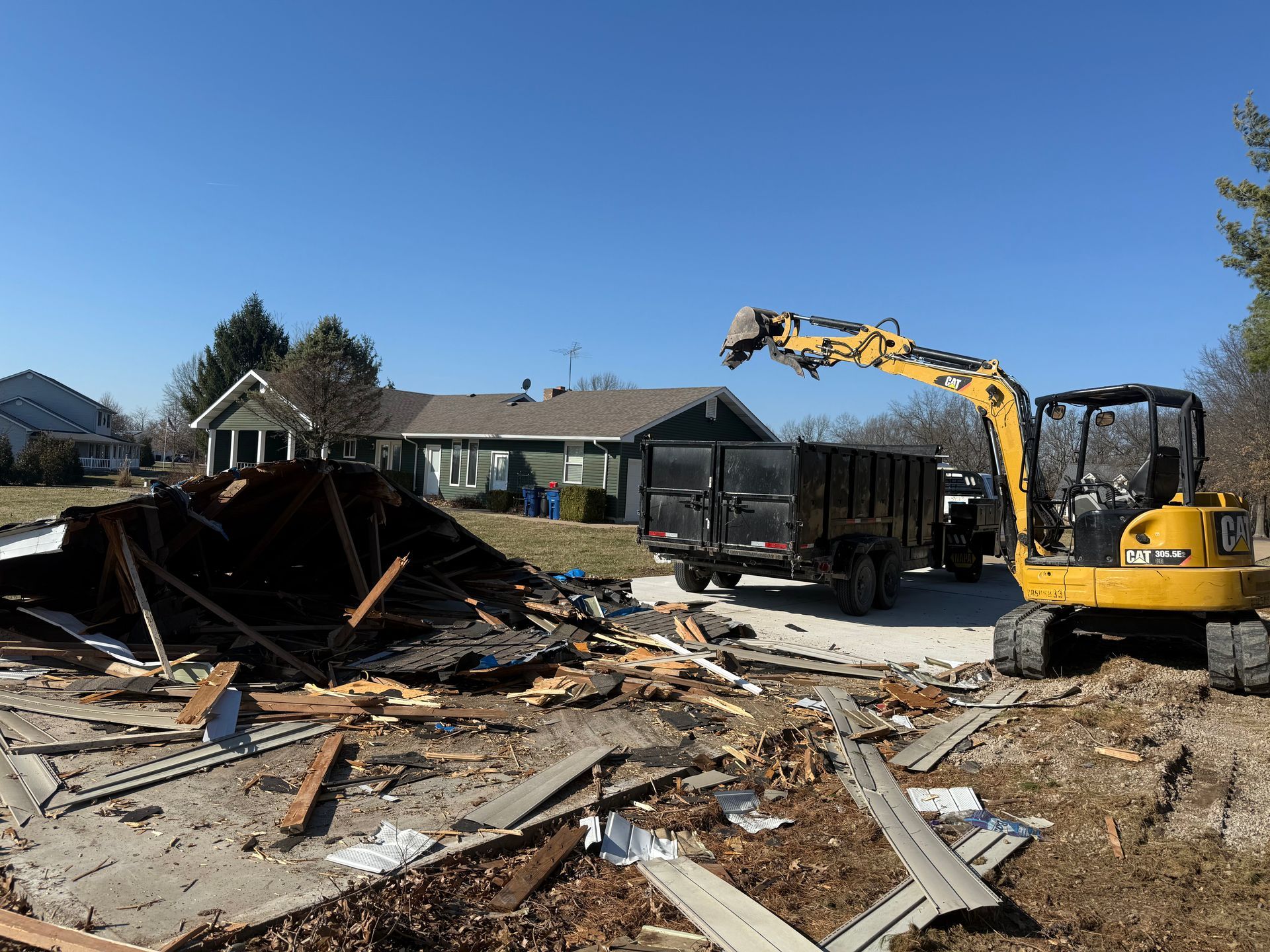 Yellow excavator clearing debris from a collapsed house in a residential neighborhood