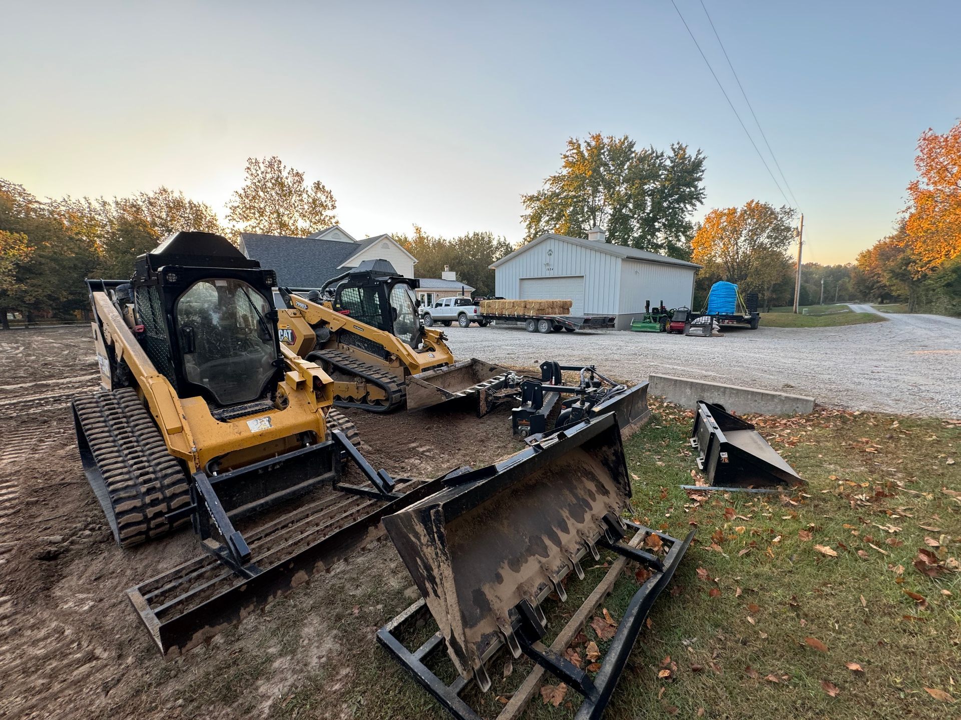 Two yellow skid-steer loaders with attachments parked on a gravel lot near a white barn at sunset