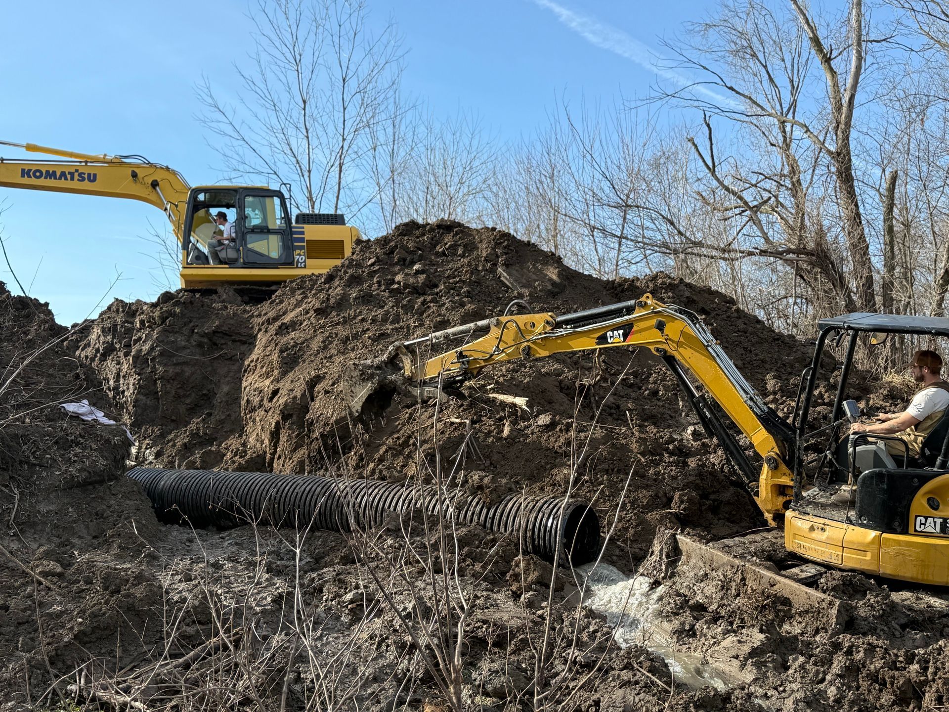 Yellow excavator clearing dirt around a large black pipe at a muddy construction site