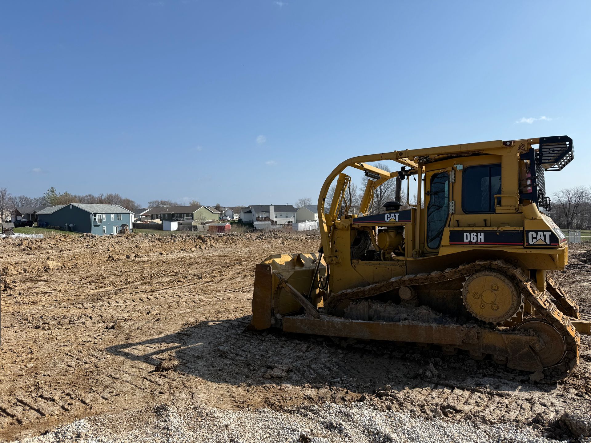 Yellow bulldozer on a dirt lot under a clear blue sky, with houses in the background.