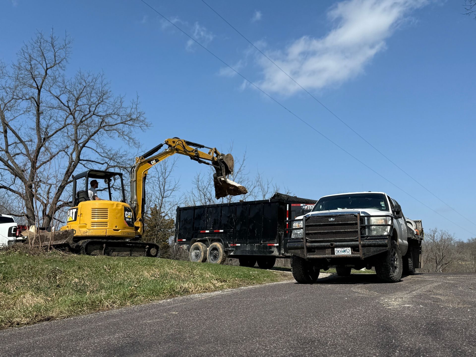Yellow excavator loading a black dump truck beside a silver pickup on a grassy roadside