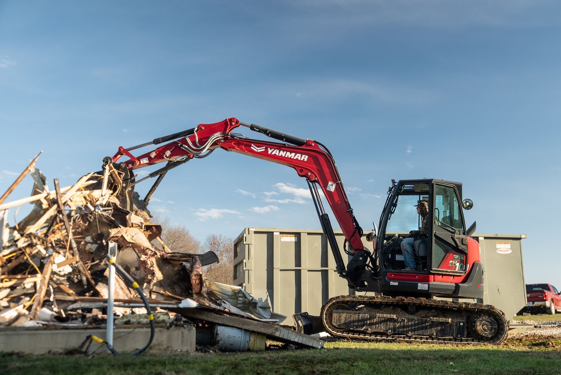 Excavator demolishing a pile of rubble beside a truck under a clear blue sky