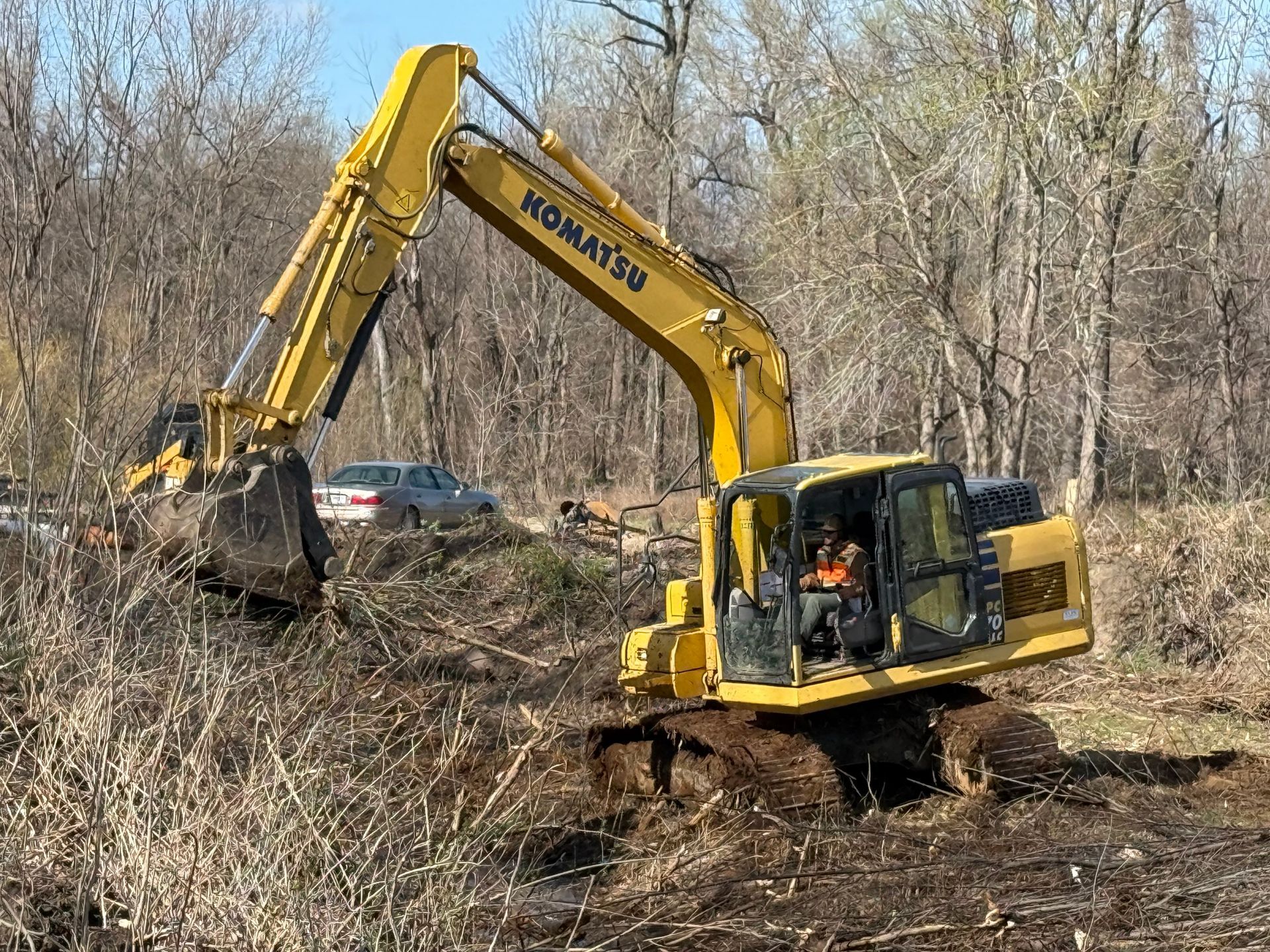 Yellow Komatsu excavator clearing brush on a wooded hillside near a parked car.