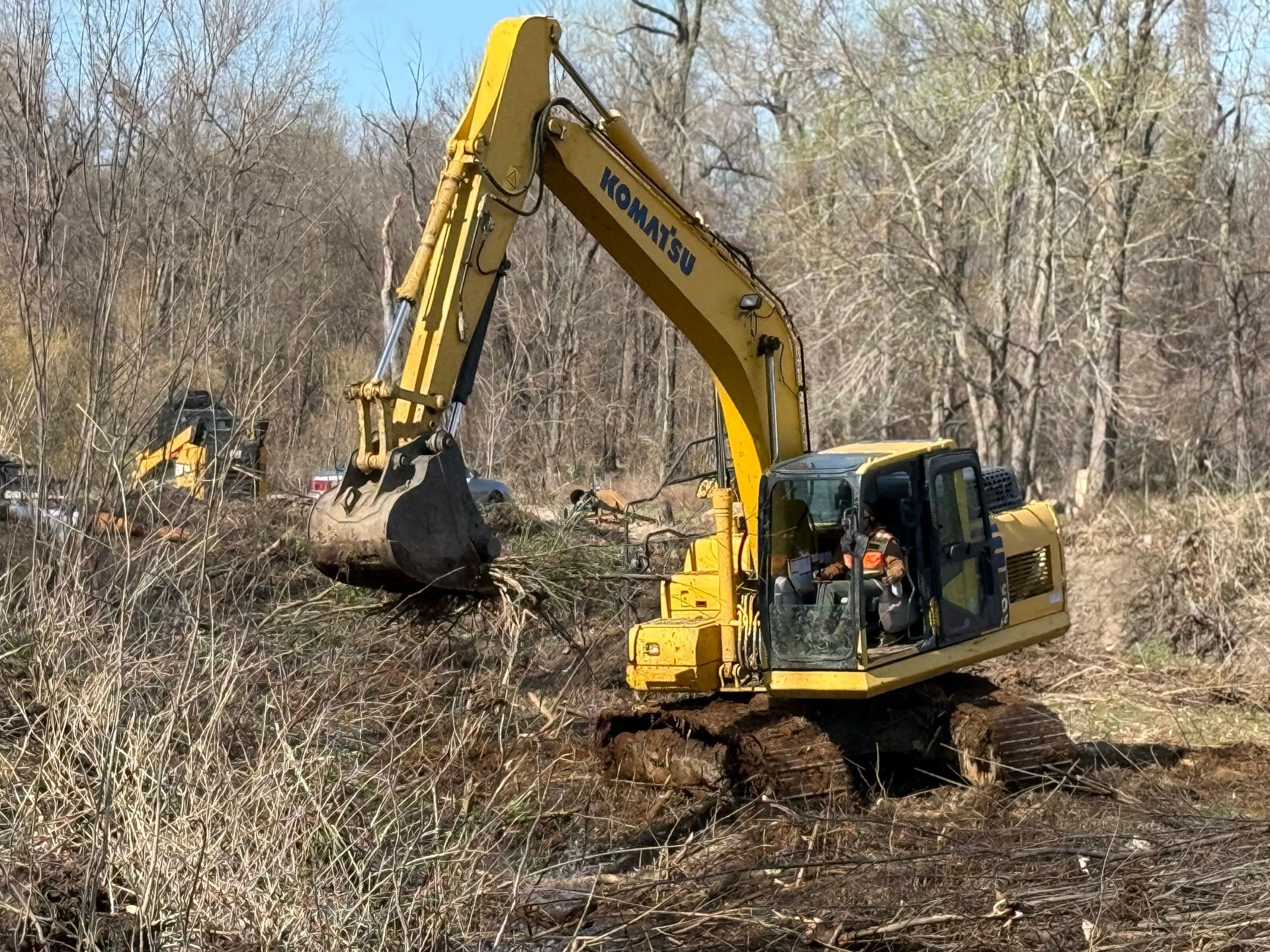 Yellow excavator clearing brush in a wooded area with a worker inside the cab.