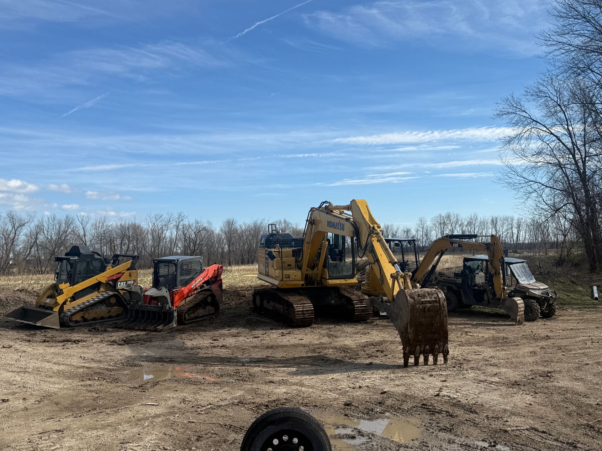 Three excavators and a loader parked in a dirt field under a blue sky