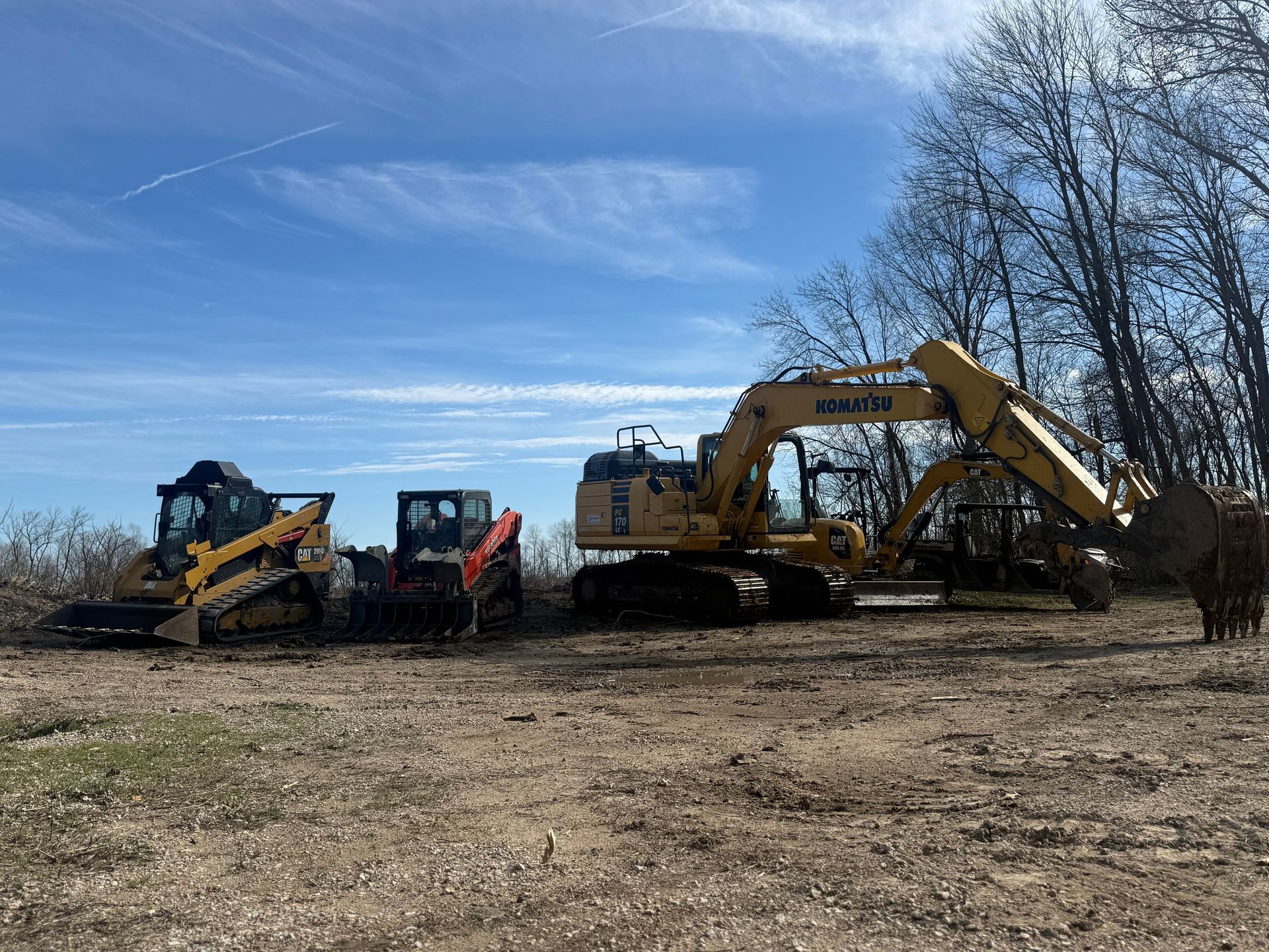 Construction equipment lined up on a dirt field under a blue sky, including an excavator and skid steer.
