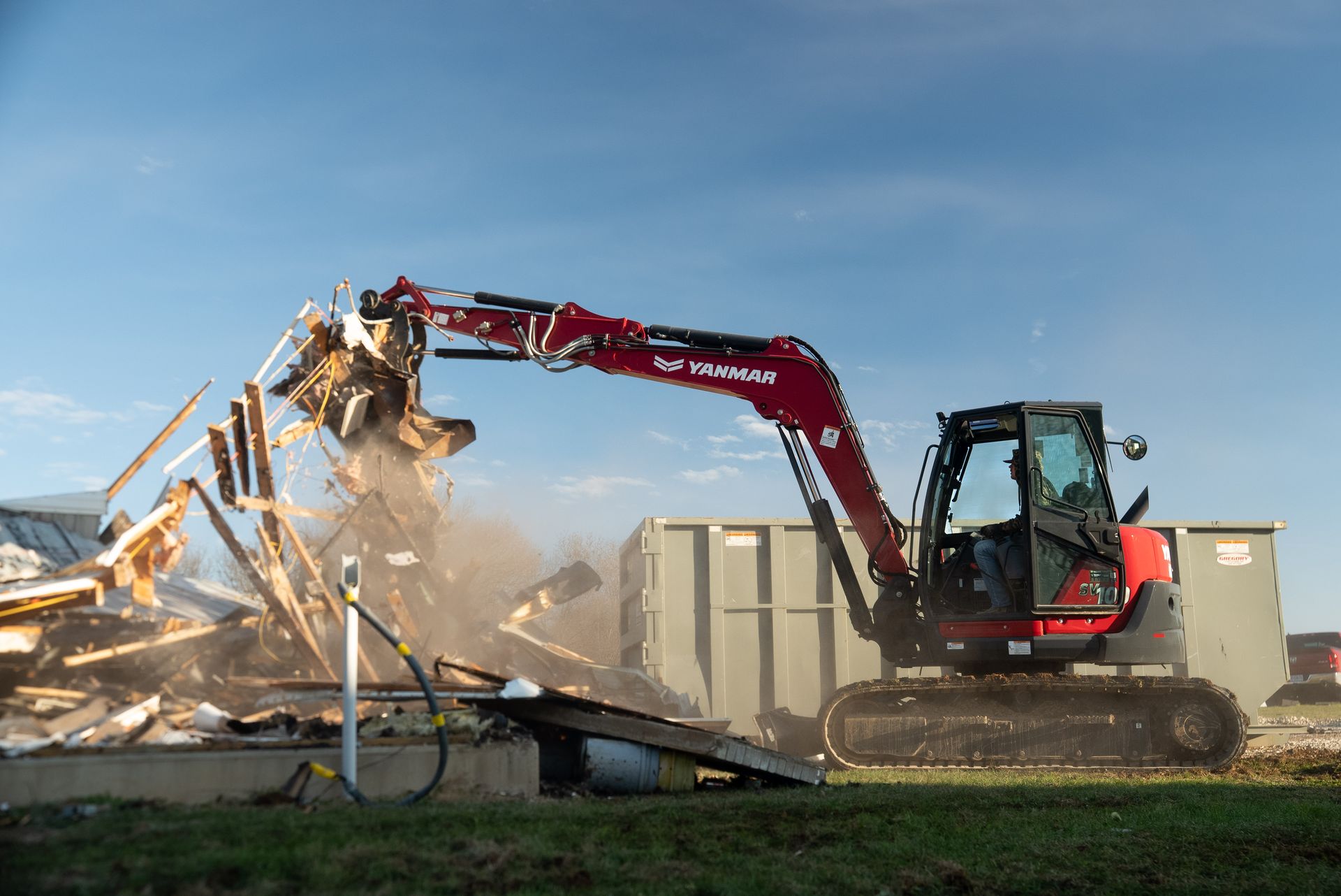 Red excavator demolishing a building, debris and dust flying under a blue sky.