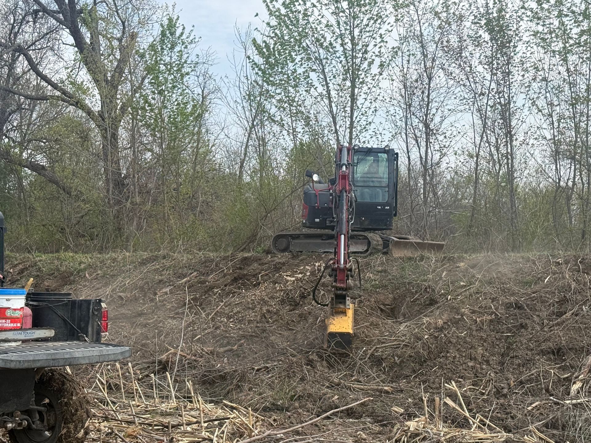 Excavator clearing brush on a wooded hillside beside a truck and equipment