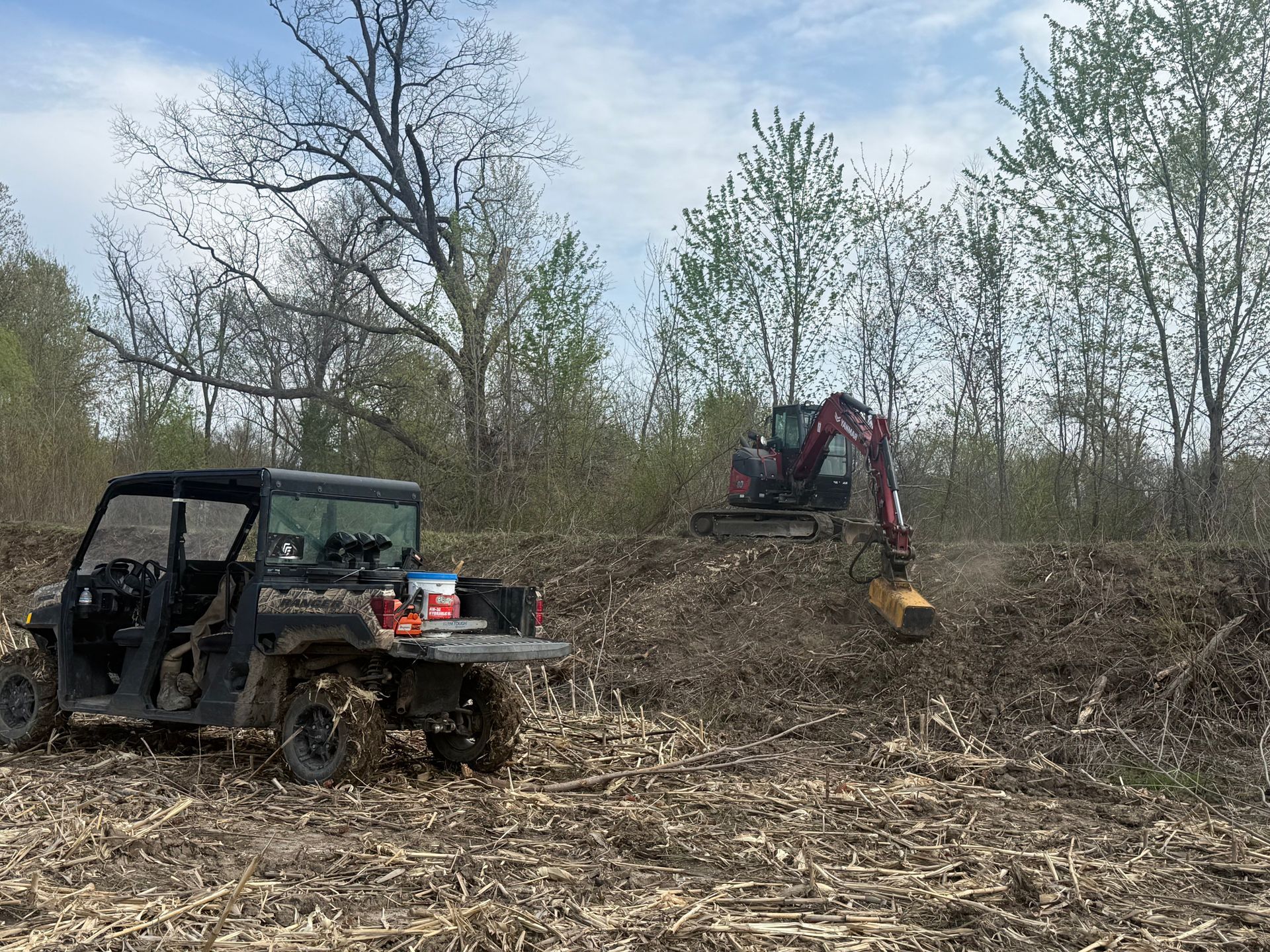 ATV and excavator clearing brush in a wooded area under a cloudy sky