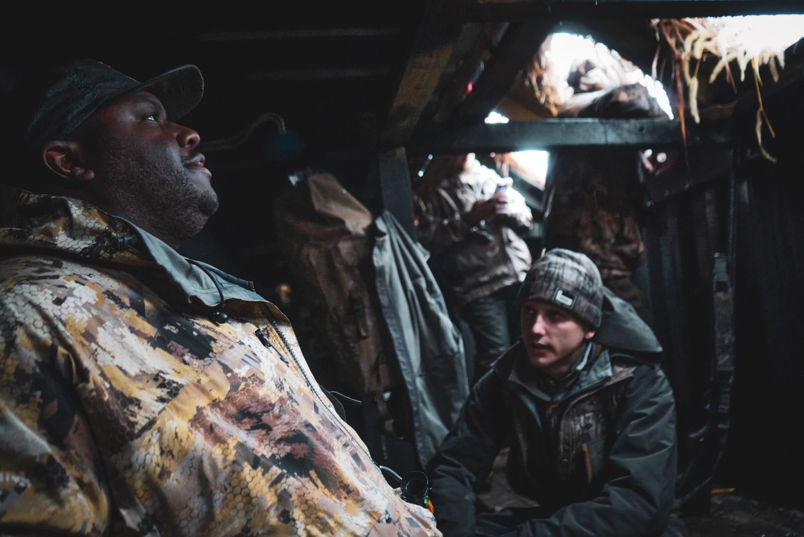 Two people in heavy winter gear inside a dark shelter, one seated and speaking, one standing nearby