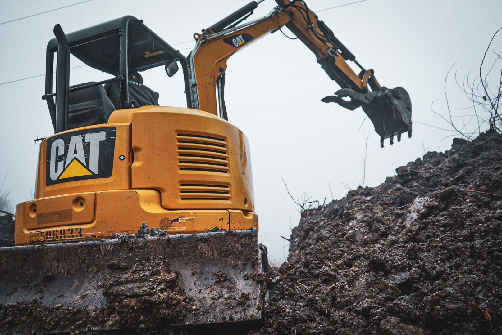Yellow CAT excavator on a muddy slope, arm raised over a pile of dirt in a gray outdoor site