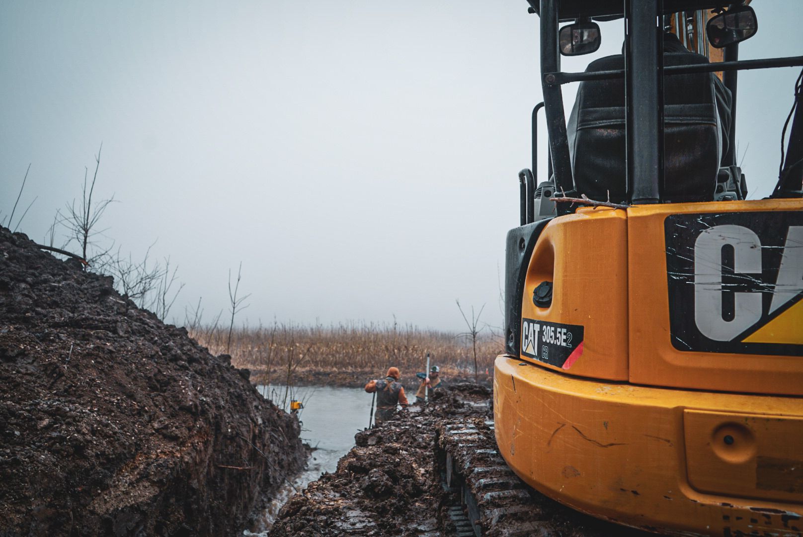 Orange CAT excavator beside a muddy trench near water under a gray sky