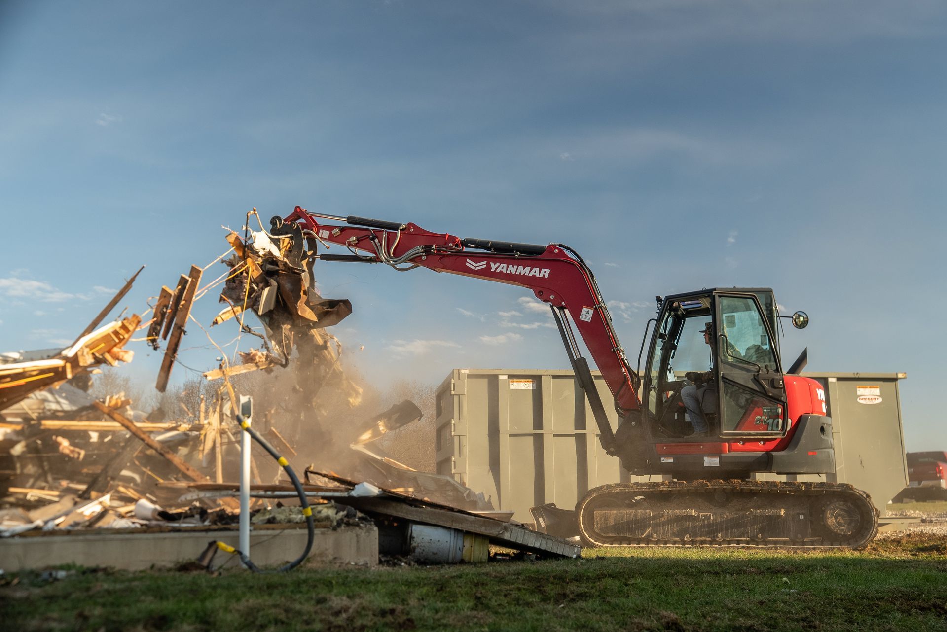 Red excavator demolishing a building, raising dust in a rural field at sunset.
