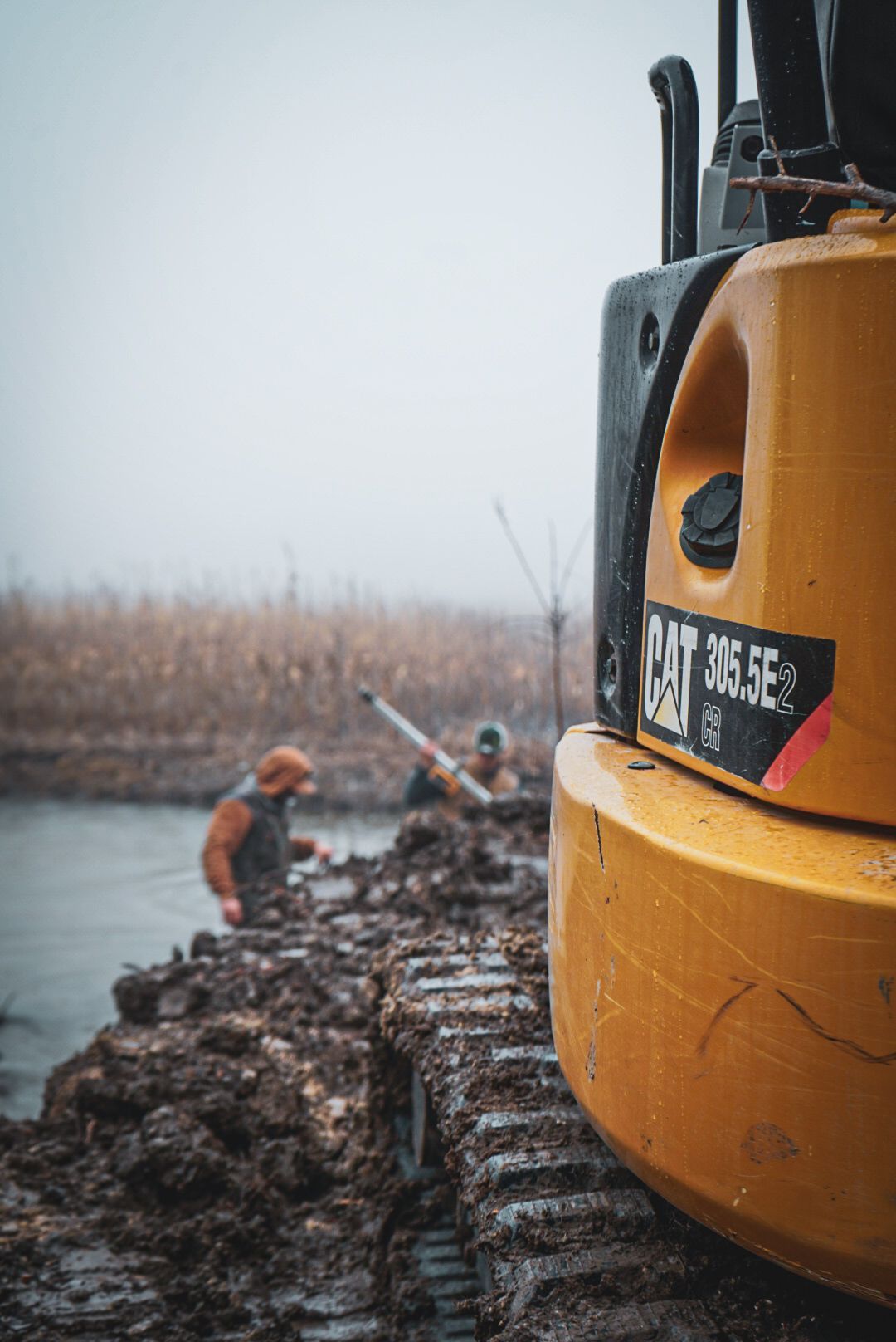 Orange excavator beside muddy trench with two workers in the background on an overcast day