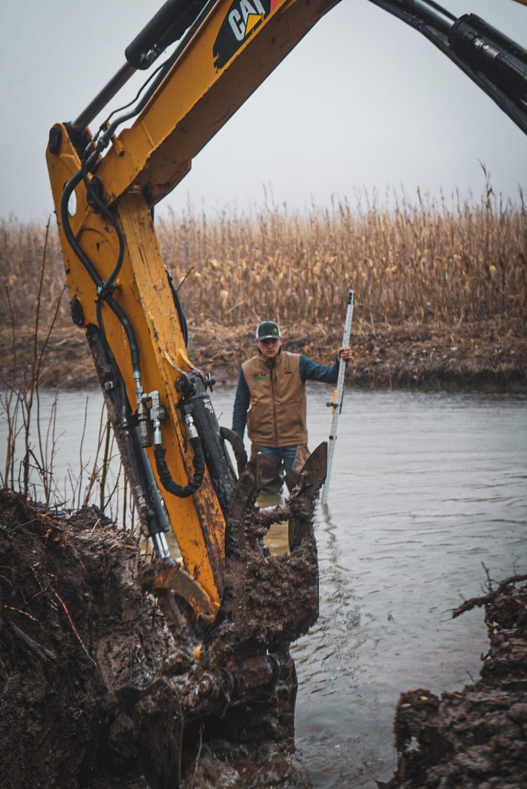 Excavator bucket at a muddy waterway, with a worker in chest waders holding a tool beside it