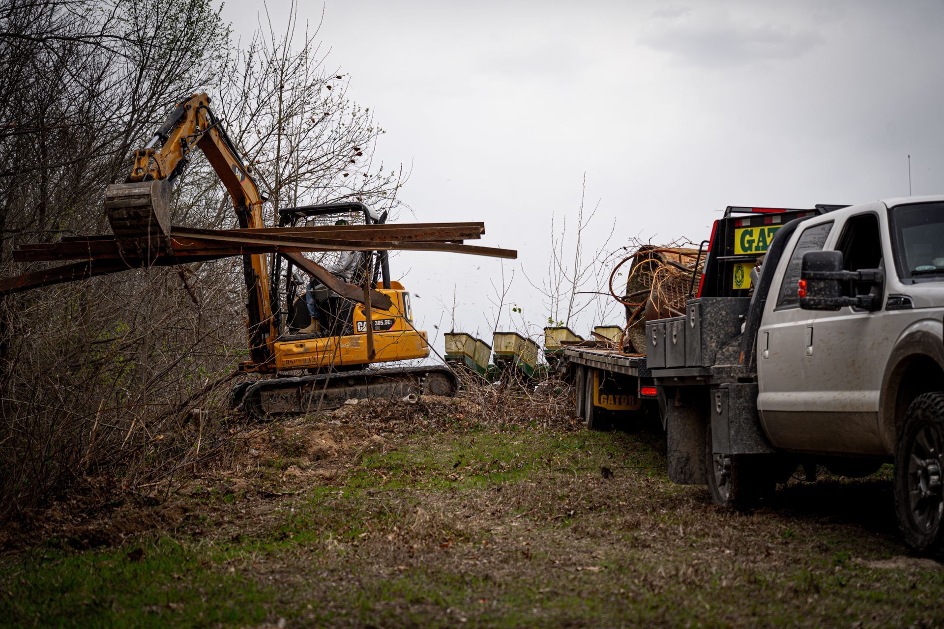 Excavator and pickup truck working at a muddy roadside clearing under a gray sky