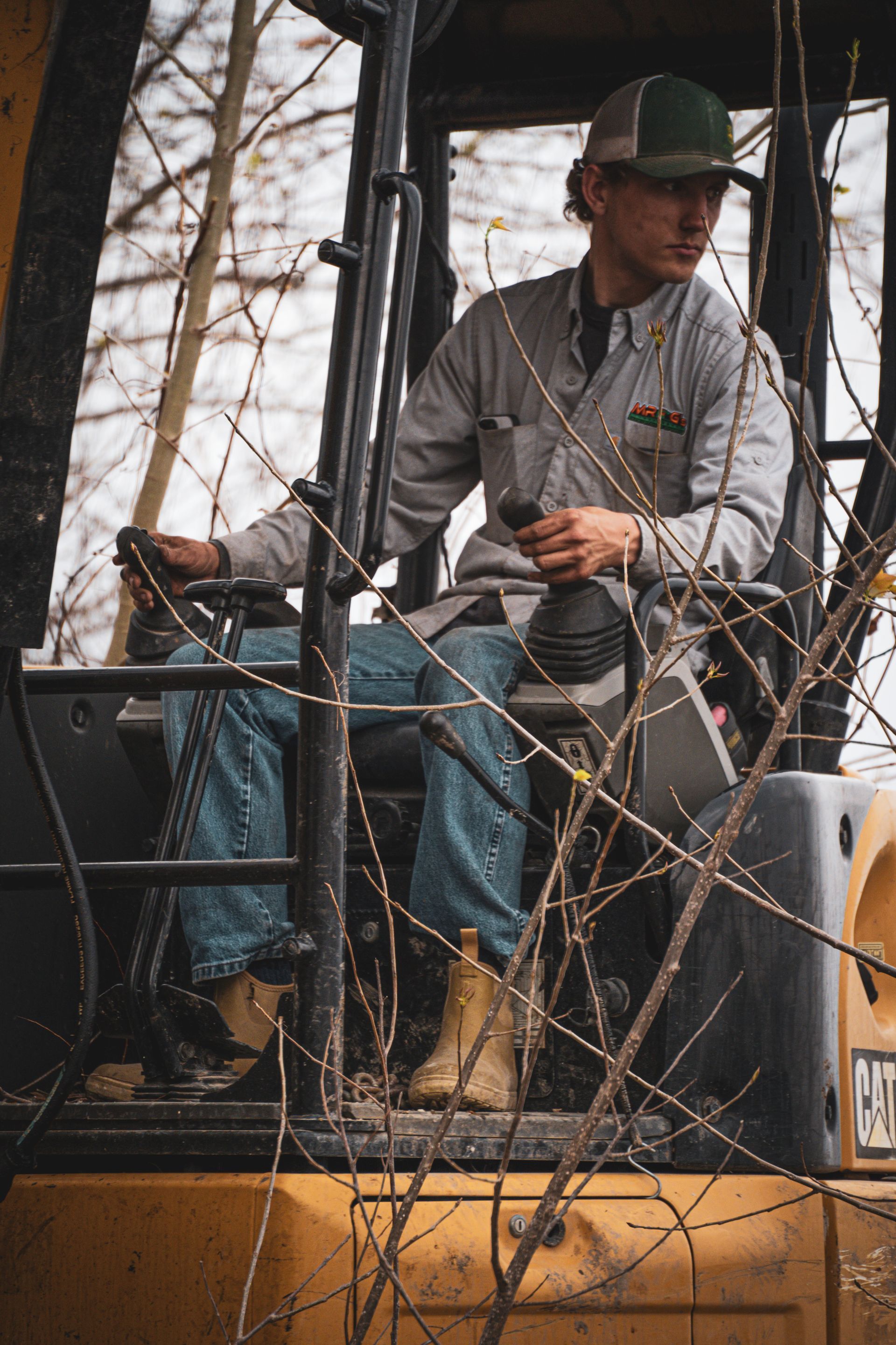 Worker operating an excavator, seated in the cab among tree branches.