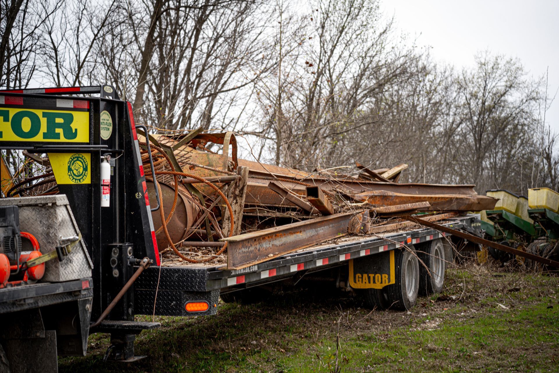 Dump trailer loaded with scrap metal and debris in a wooded area, with a tow vehicle at left.