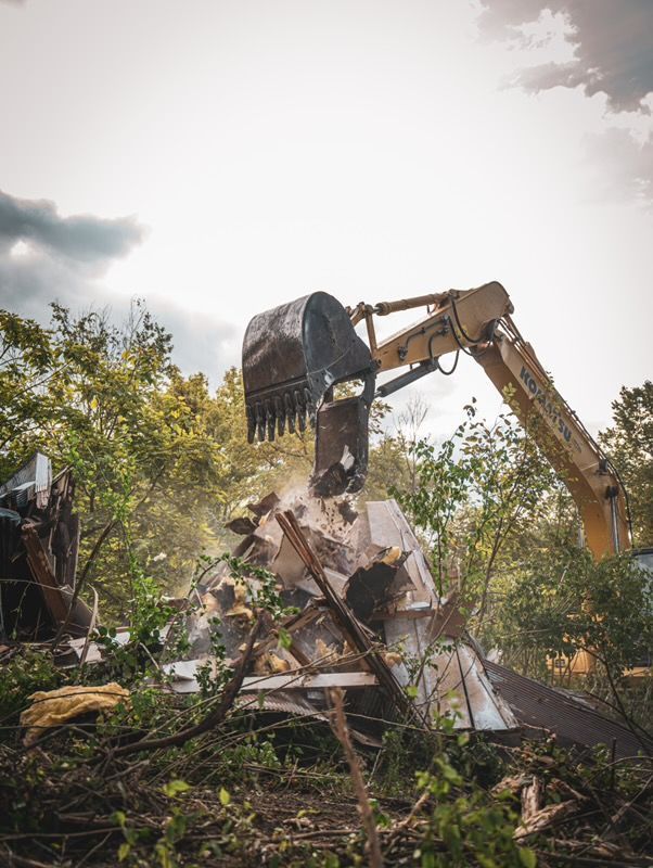 Excavator demolishing a ruined structure amid overgrown greenery and dust