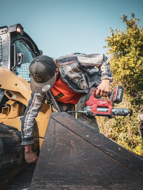 Worker using a red chainsaw cuts a dark board beside a yellow excavator outdoors