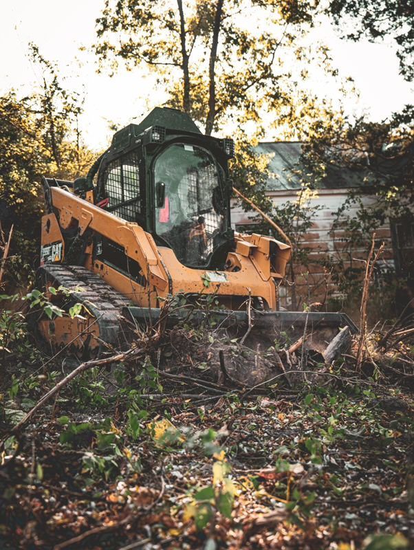 Orange excavator clearing brush in a wooded area near a house