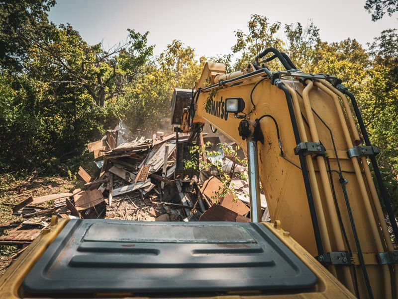Yellow excavator demolishing a pile of debris in a wooded outdoor area