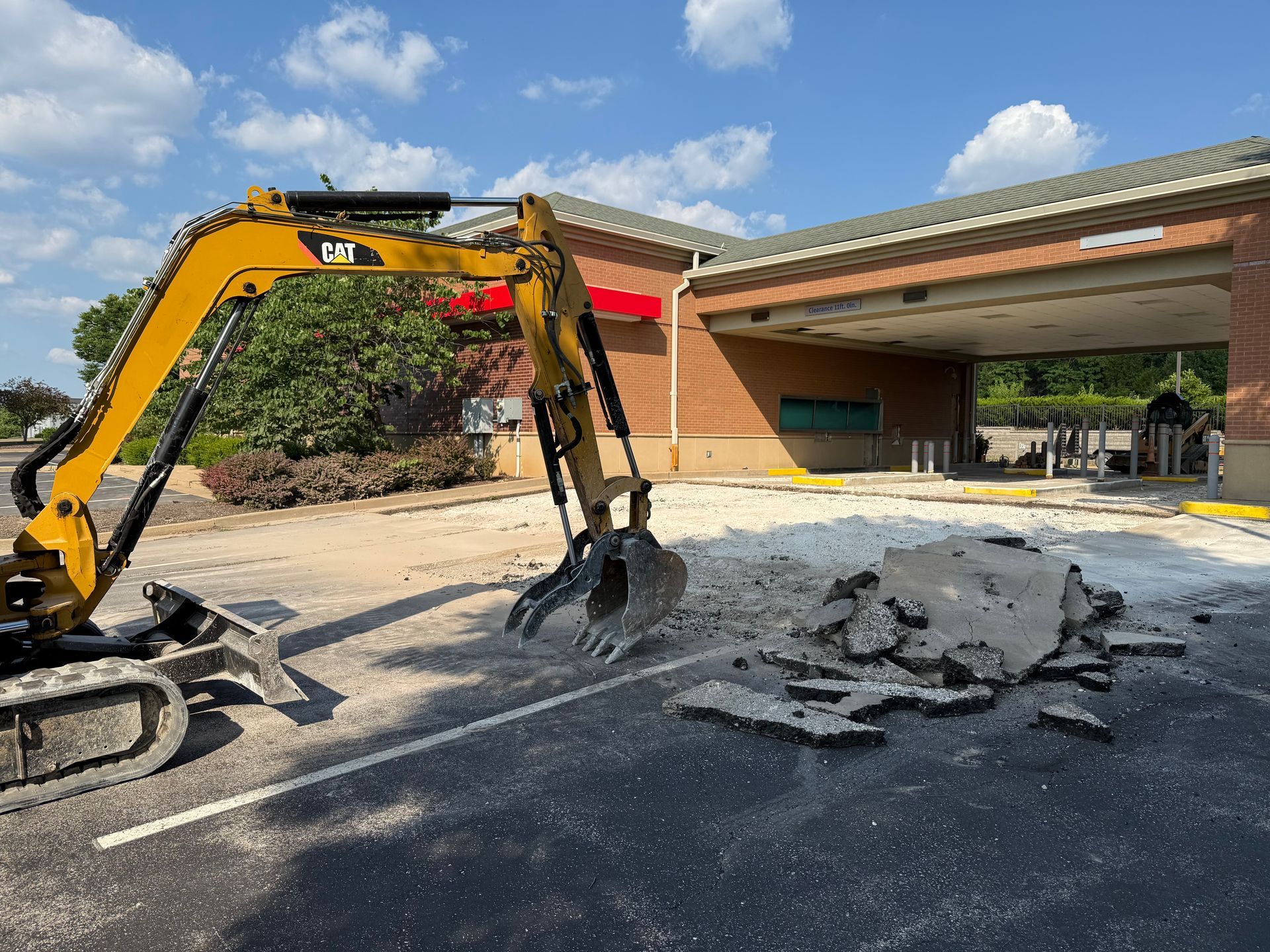 Yellow excavator breaking up concrete in a driveway beside a garage with debris piled nearby