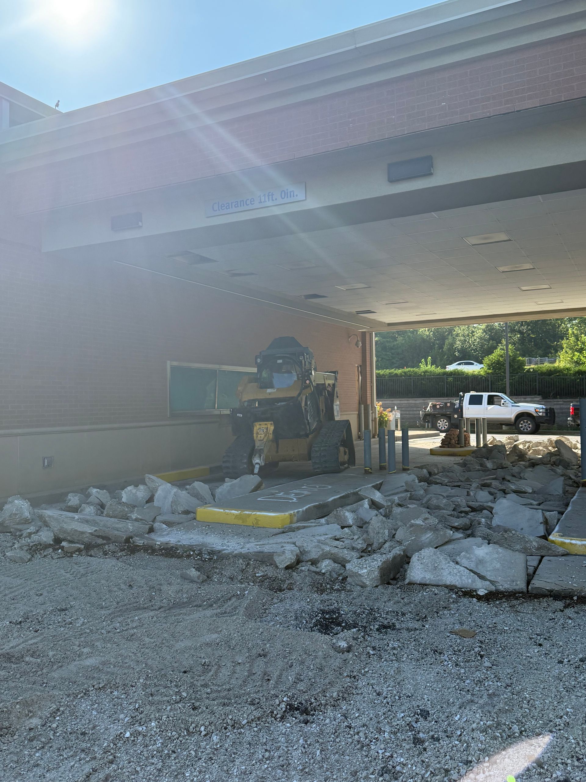 Construction equipment clearing rubble under a covered building entrance in bright sunlight