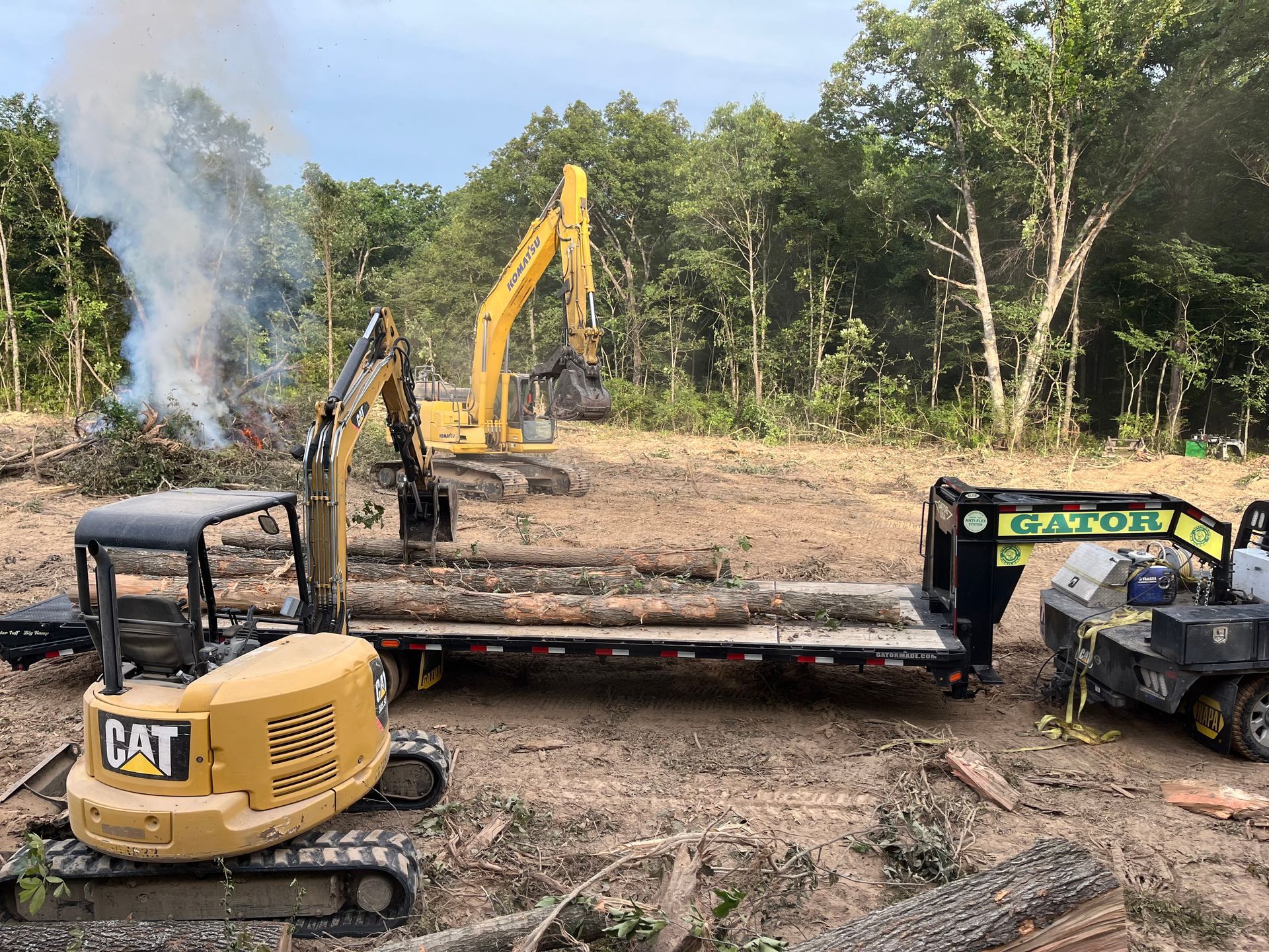 Excavators and a flatbed trailer at a muddy construction site with smoke rising nearby.