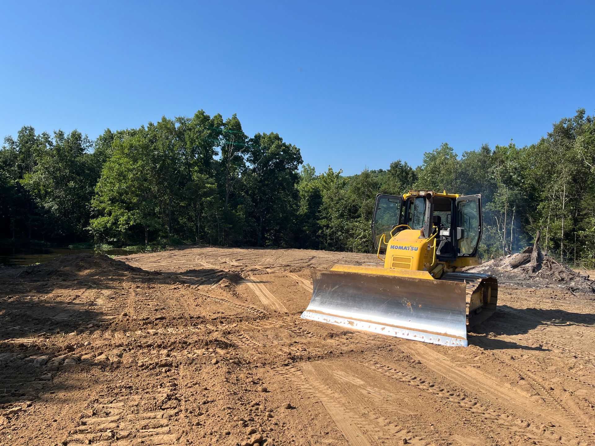 Yellow bulldozer grading a dirt lot beside trees under a clear blue sky
