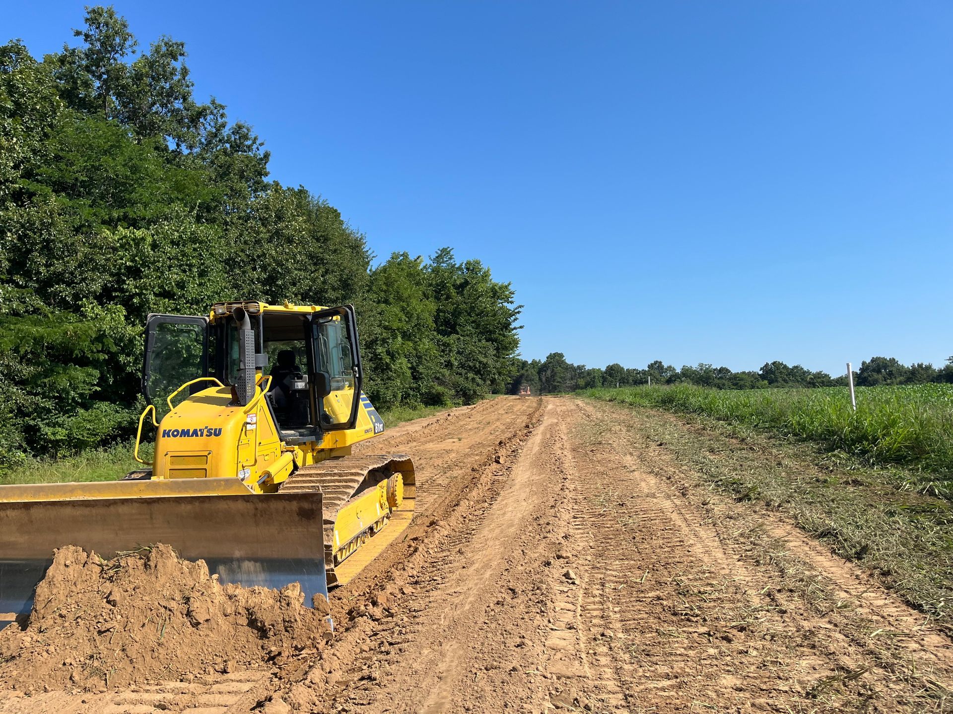 Yellow bulldozer grading a dirt road beside green fields under a clear blue sky