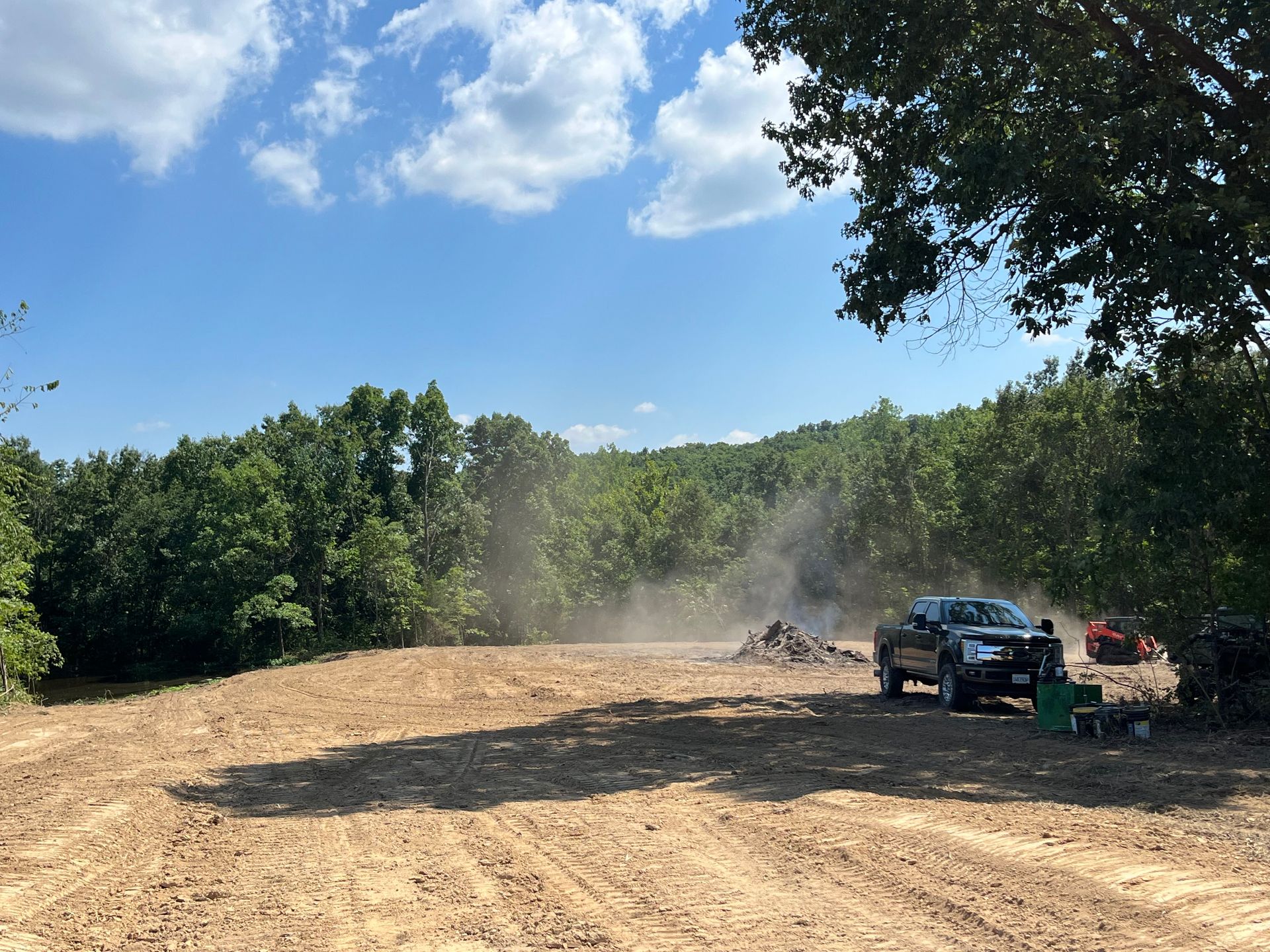 Open dirt clearing with dust, parked vehicles, and people near a tree line under a blue sky