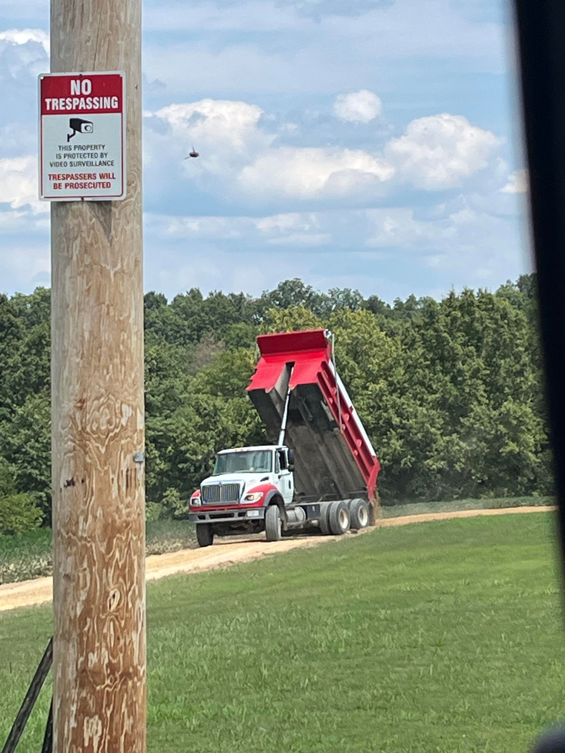 Red dump truck hauling dirt on a rural gravel road beside a grassy field and trees