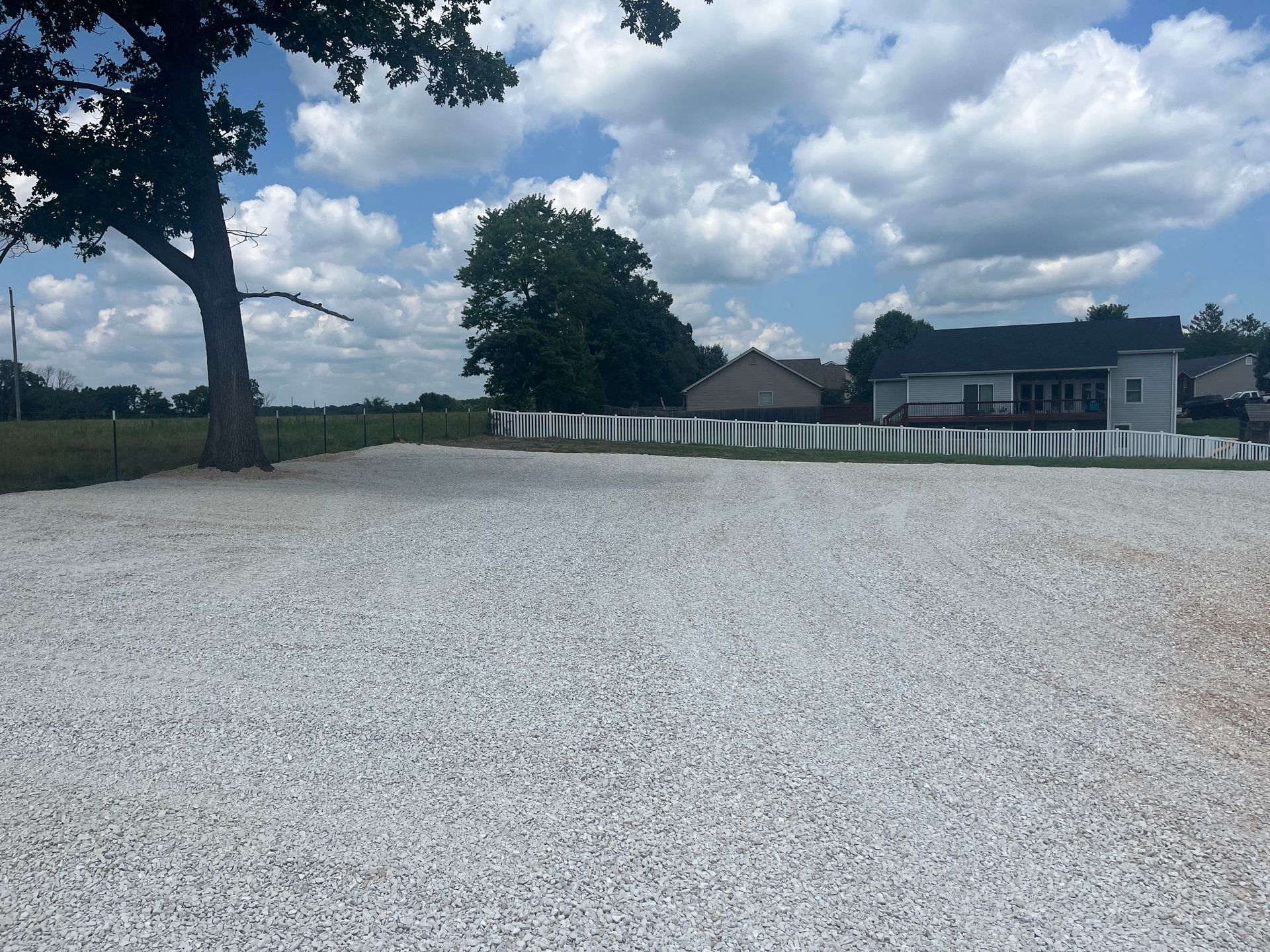 Gravel courtyard with a tree, white fence, and house under a partly cloudy sky