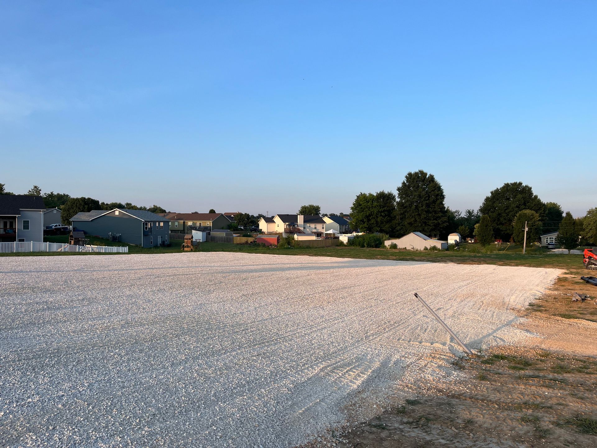 Gravel lot beside trees and houses under a clear blue sky at dusk