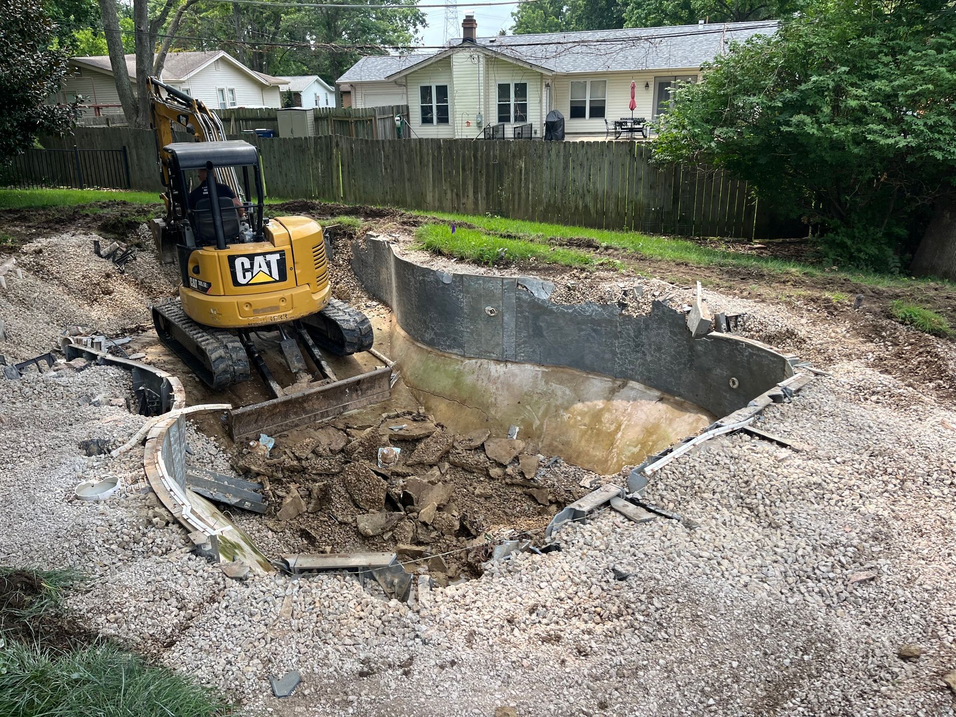Yellow CAT excavator digging a large pit in a gravel lot beside a residential fence and houses