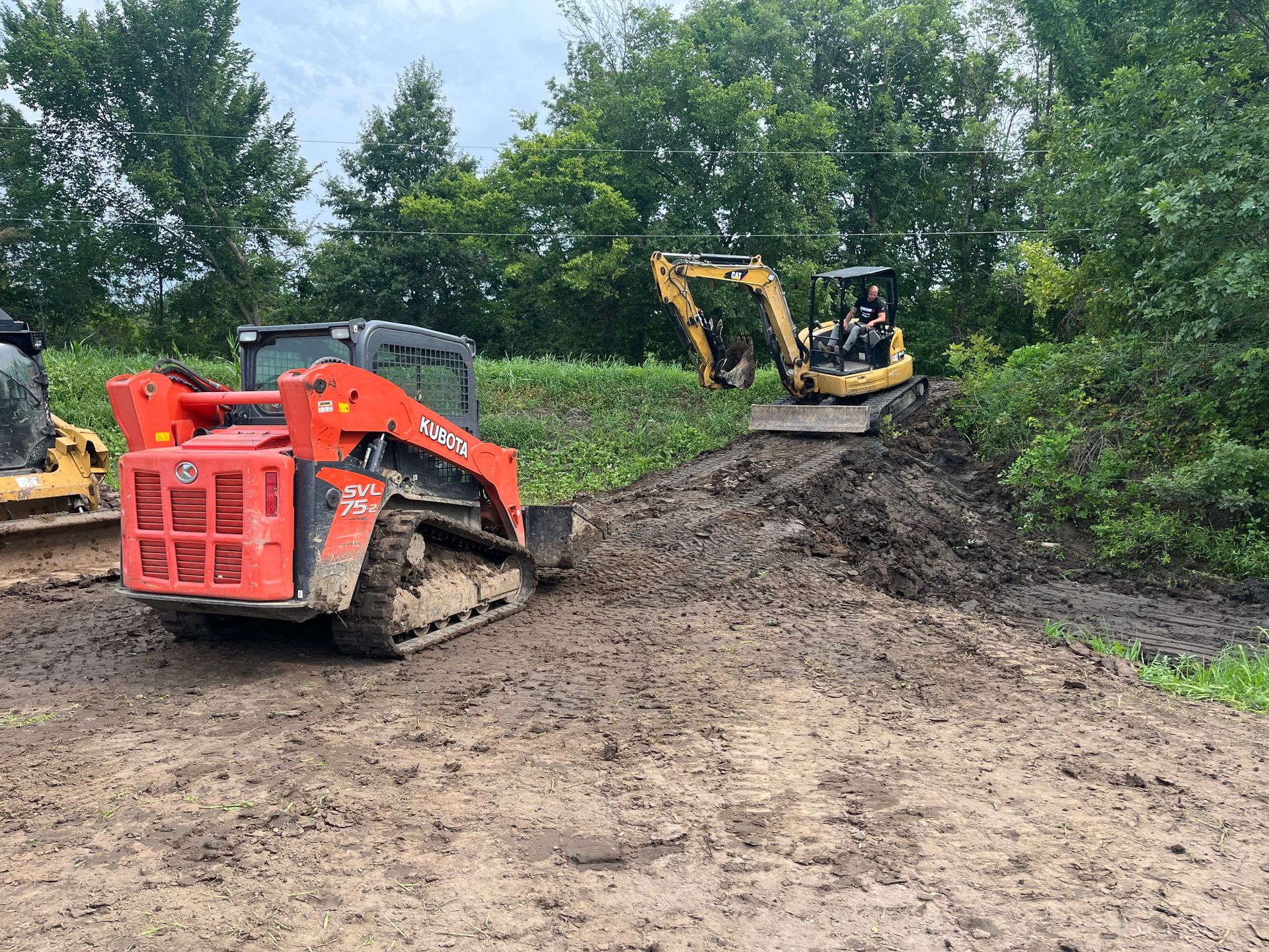 Orange skid steer and yellow excavator working on a dirt pile in a wooded construction site.