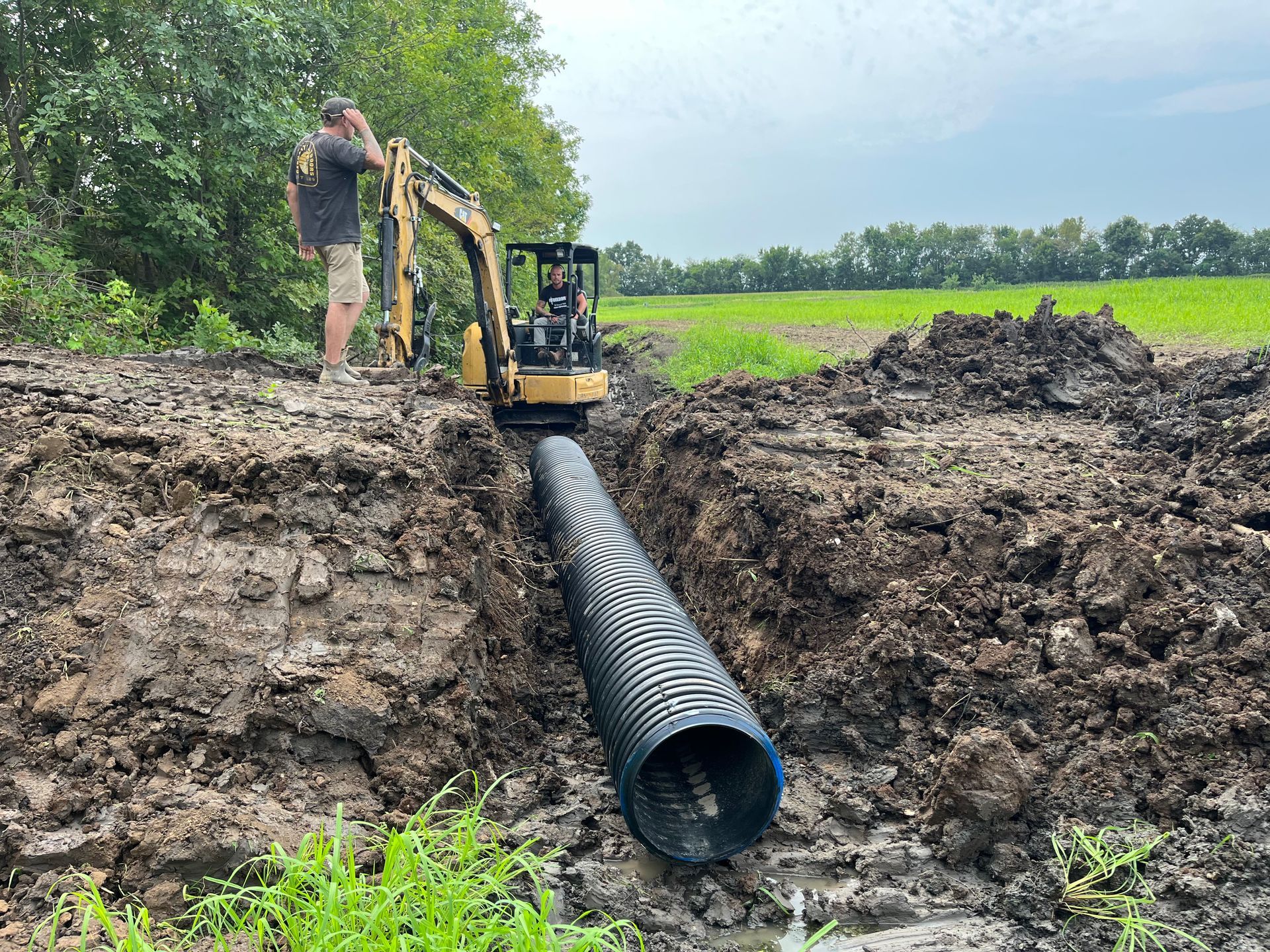 Excavator installing a large black drainage pipe in a muddy trench beside a green field