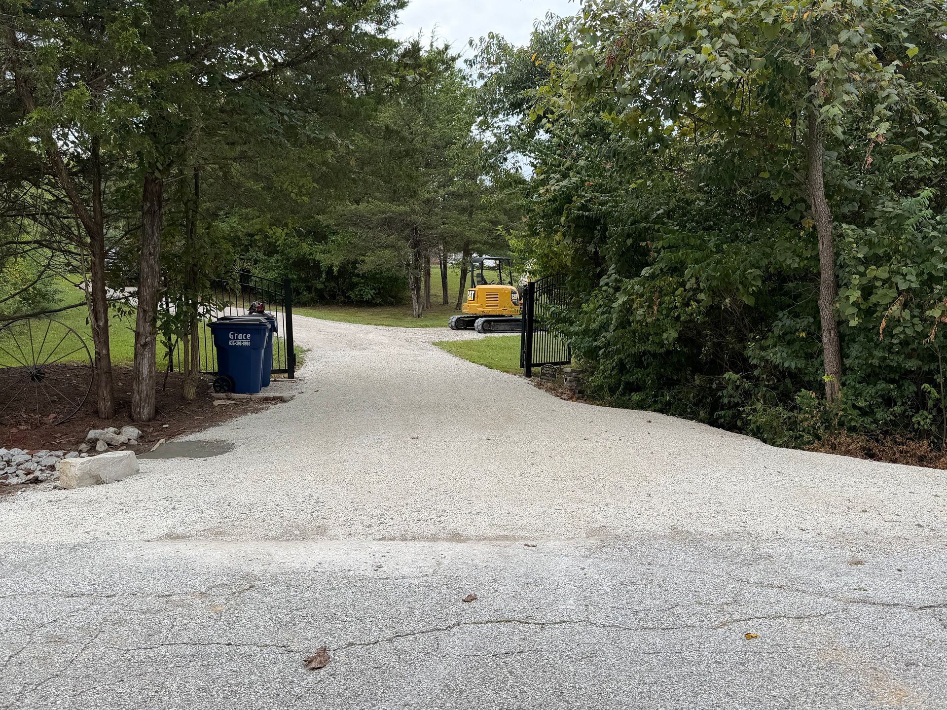 Gravel driveway lined with trees and hedges, with blue bins on the left and a yellow vehicle in the distance.