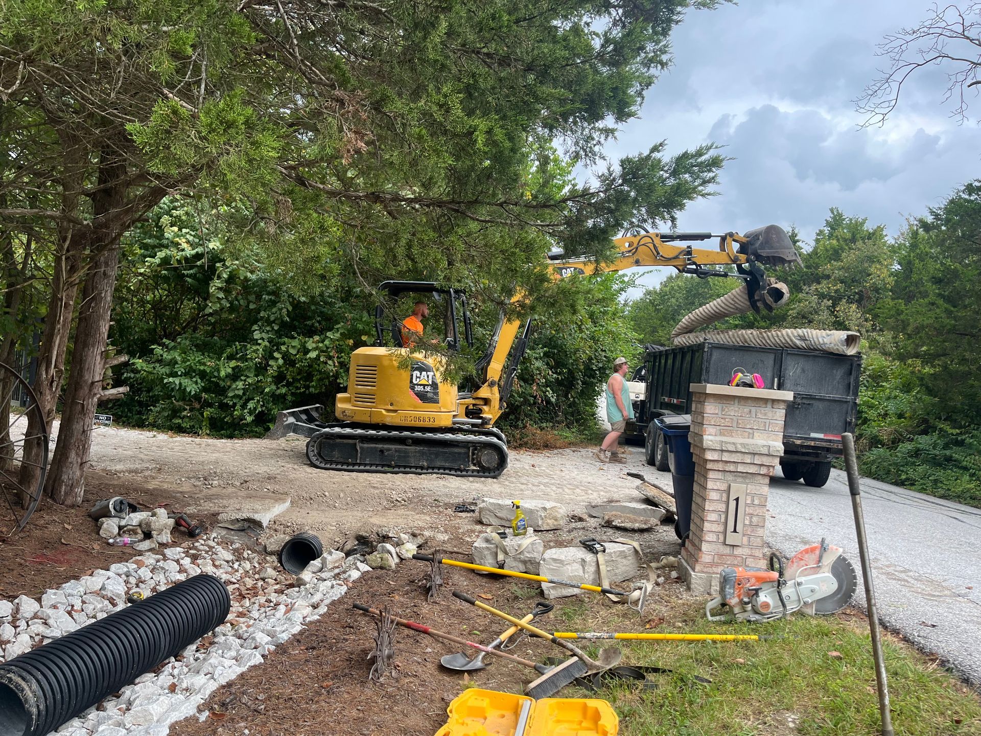 Construction crew operating a yellow excavator on a gravel driveway beside a wooded road.